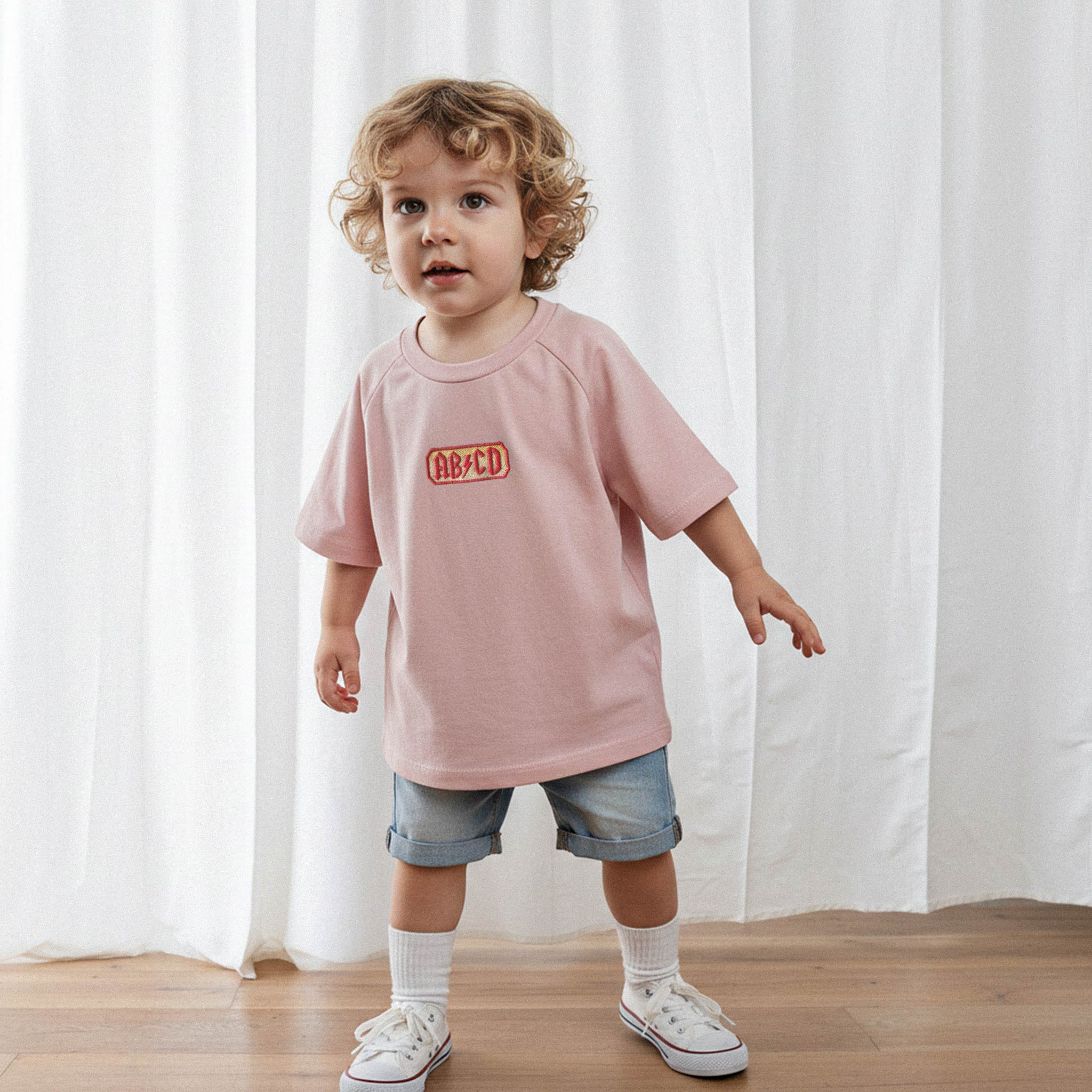 Jean şort ve gül kurusu 'AB/CD' nakışlı tişört giyen kıvırcık saçlı küçük erkek bebek, meraklı bir ifadeyle kameraya bakıyor.