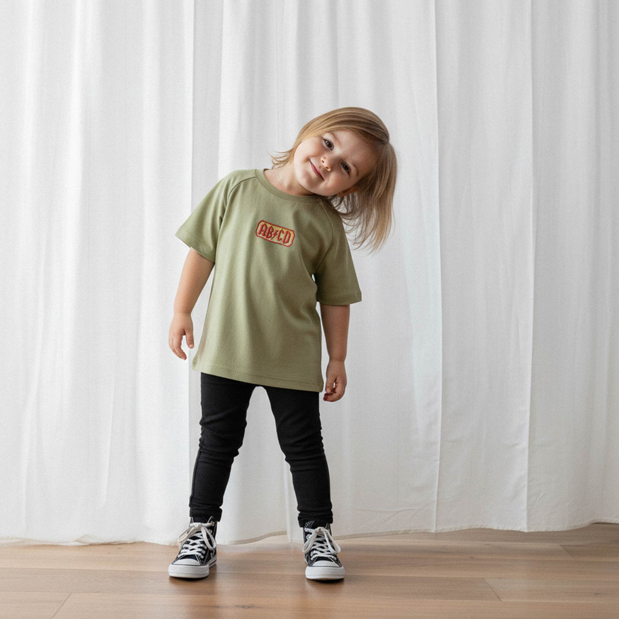 Siyah tayt giyen sevimli kız bebek, üzerinde haki yeşili renkli 'AB/CD' nakışlı oversize tişört ile başını yana eğerek gülümsüyor.