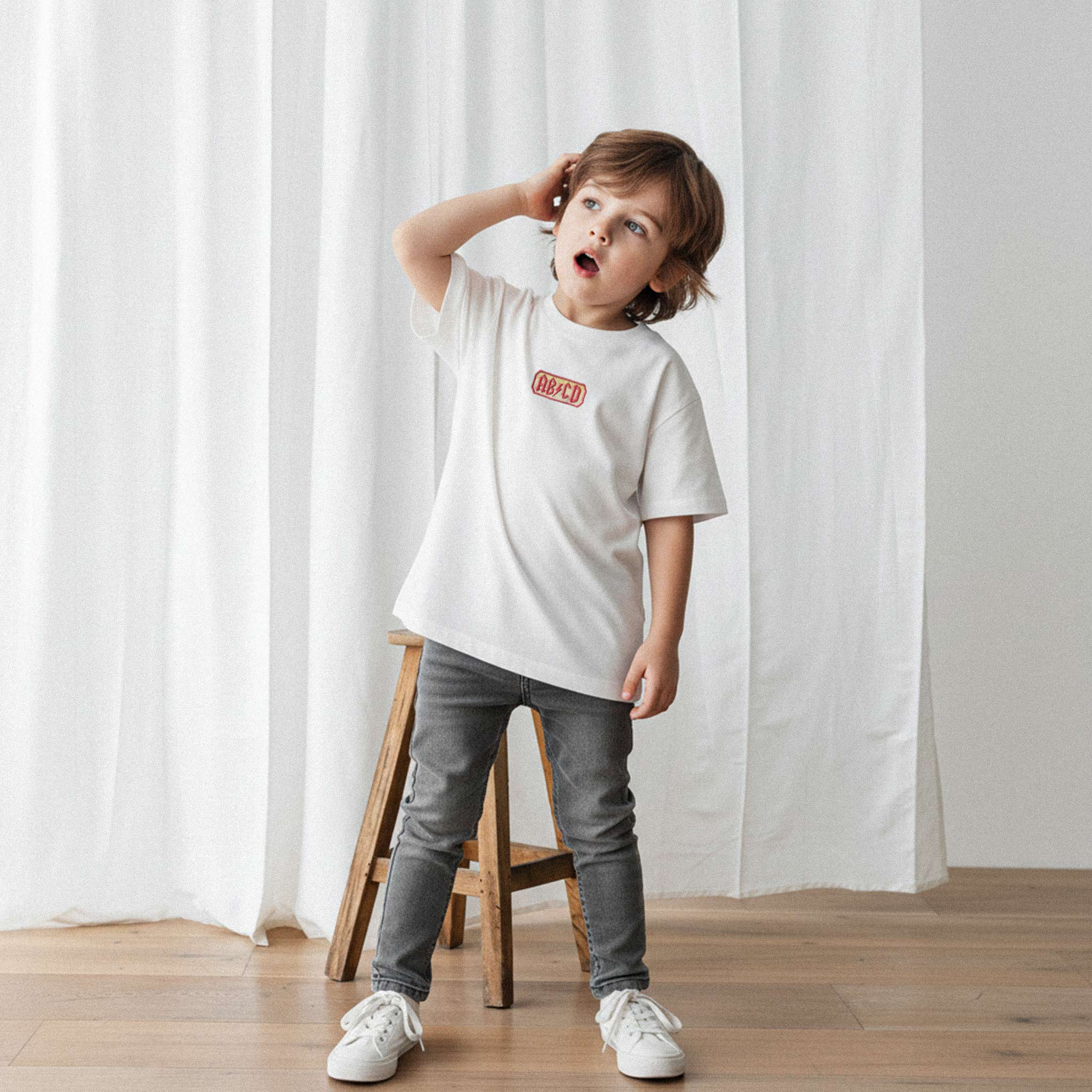 Üzerinde ekru renkli 'AB/CD' nakışlı tişört bulunan erkek çocuk, eliyle saçını düzeltirken şaşkın bir ifadeyle yukarı bakıyor.