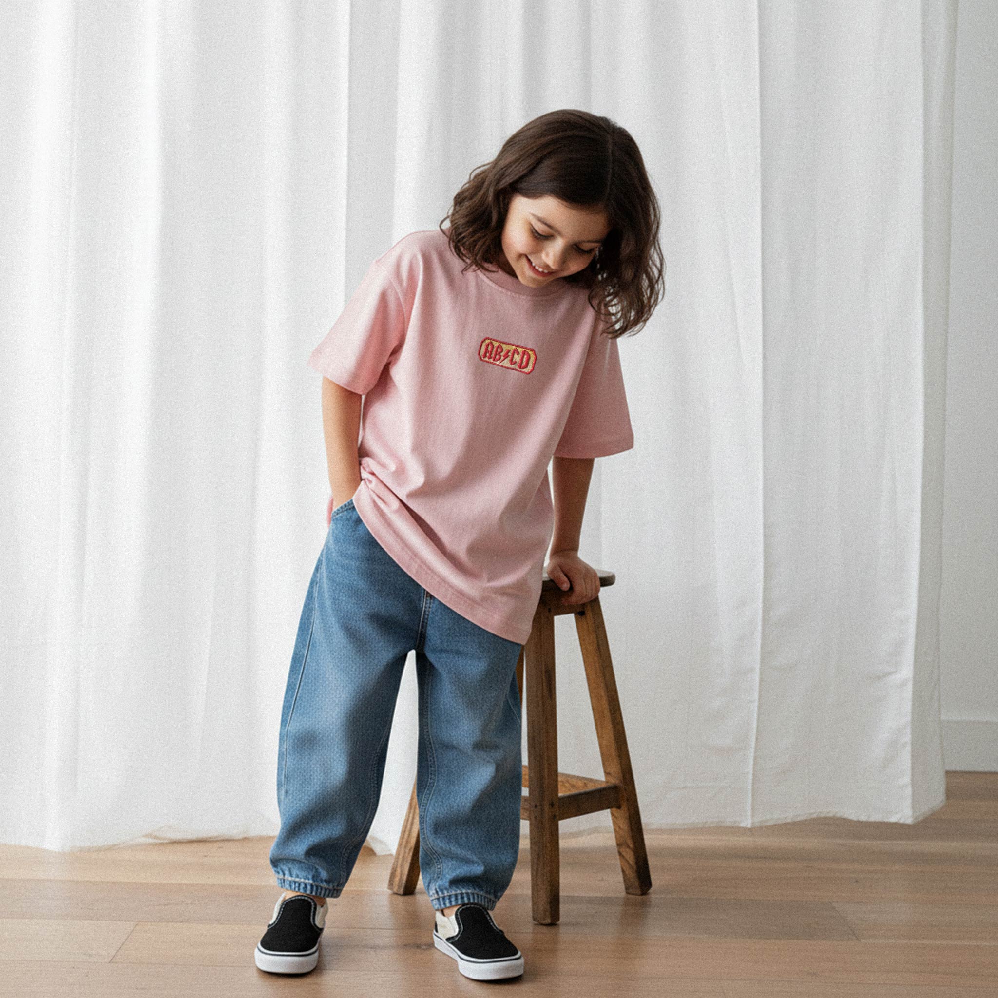 Bol paça jean pantolon ve gül kurusu 'AB/CD' nakışlı tişört giyen kız çocuk, sandalyeye dayanmış, tişörtüne bakarak gülümsüyor.