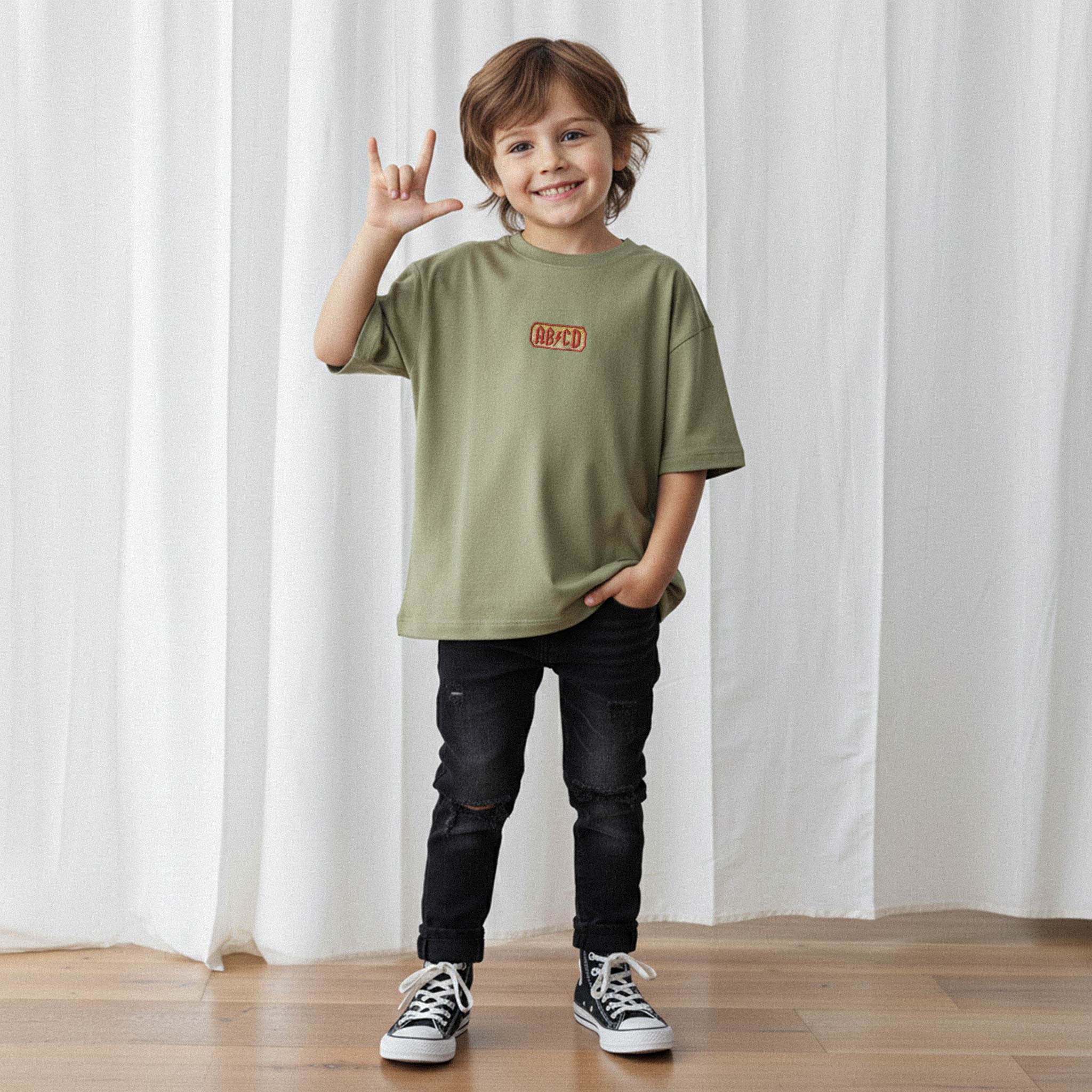 Siyah jean pantolon giyen gülümseyen erkek çocuk, üzerinde haki yeşili 'AB/CD' nakışlı oversize tişört ile rock işareti yaparak poz veriyor.