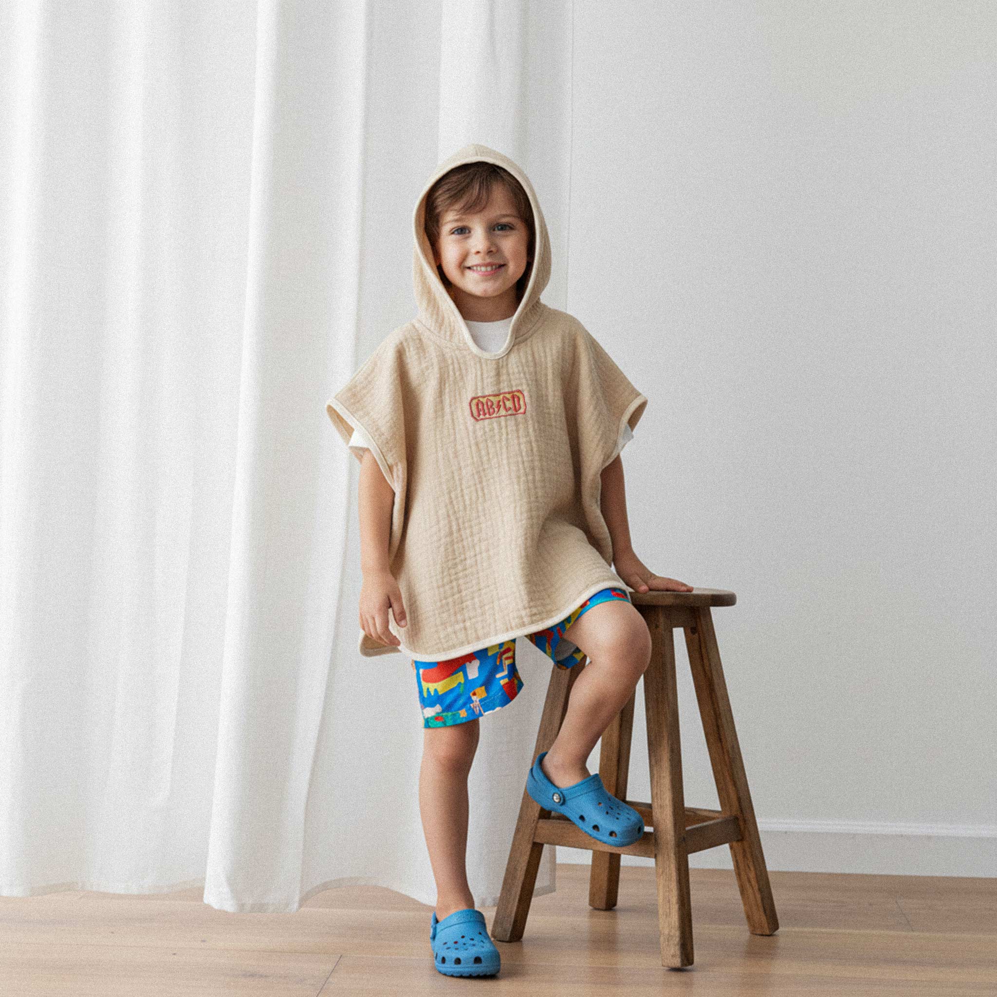 Renkli deniz şortu ve somon rengi, 'AB/CD' nakışlı kapüşonlu müslin panço giyen erkek çocuk, ahşap bir tabureye yaslanarak gülümsüyor.