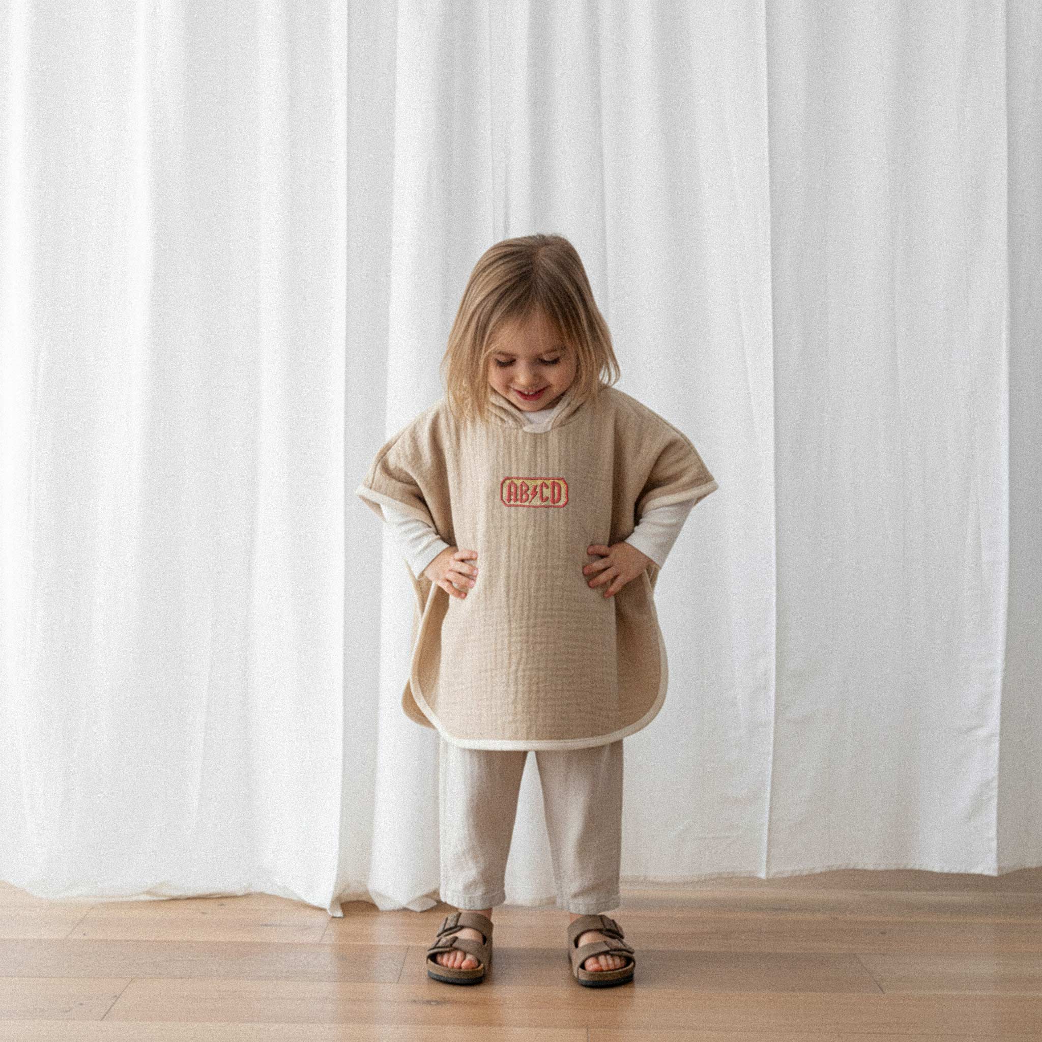 Keten pantolon giyen kız çocuk, üzerinde somon rengi, 'AB/CD' nakışlı kapüşonlu müslin panço ile elleri belinde gülümseyerek pançosuna bakıyor.