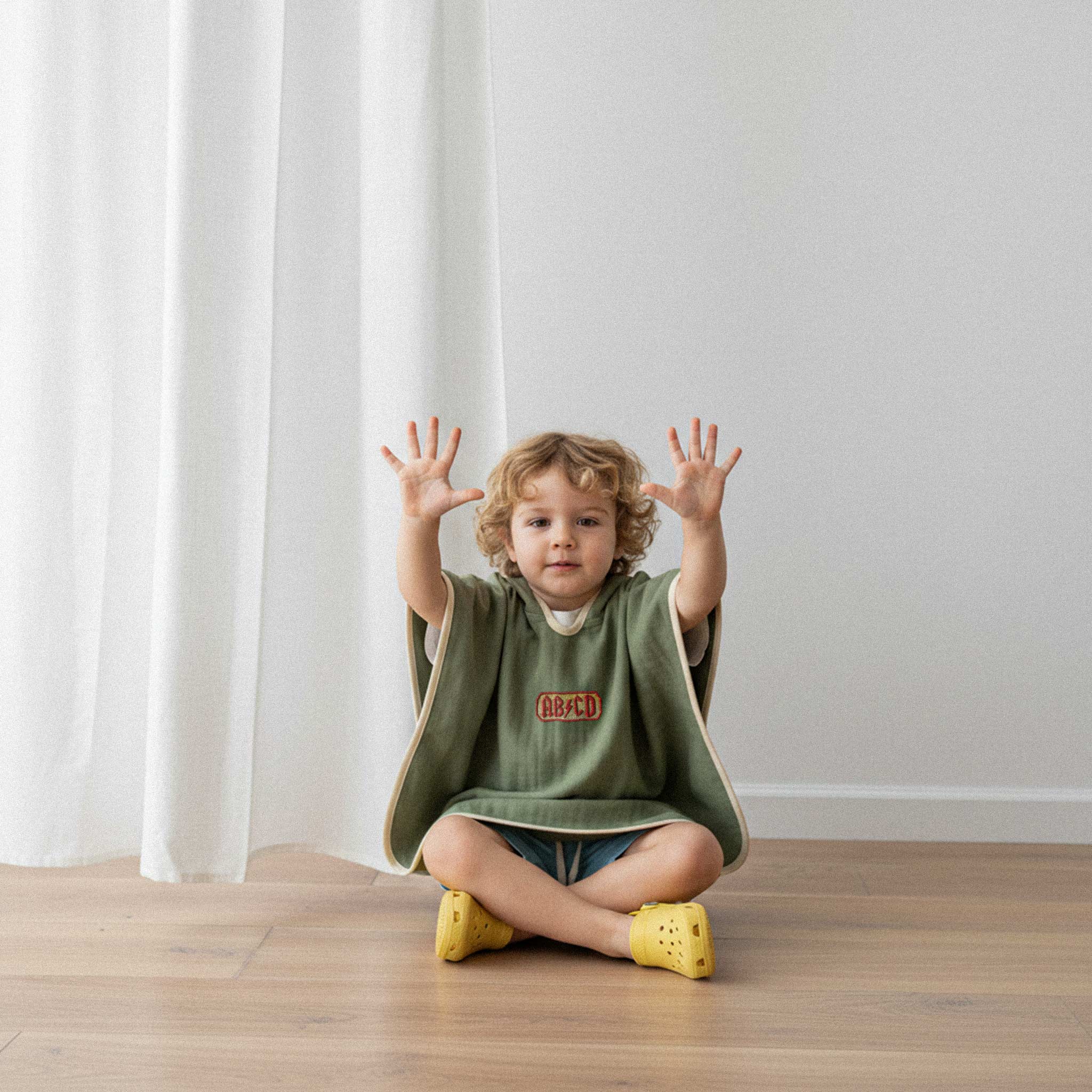 Şort giyen kıvırcık saçlı küçük erkek çocuk, üzerinde haki yeşili, 'AB/CD' nakışlı kapüşonlu müslin panço ile yerde oturarak ellerini havaya kaldırıyor.