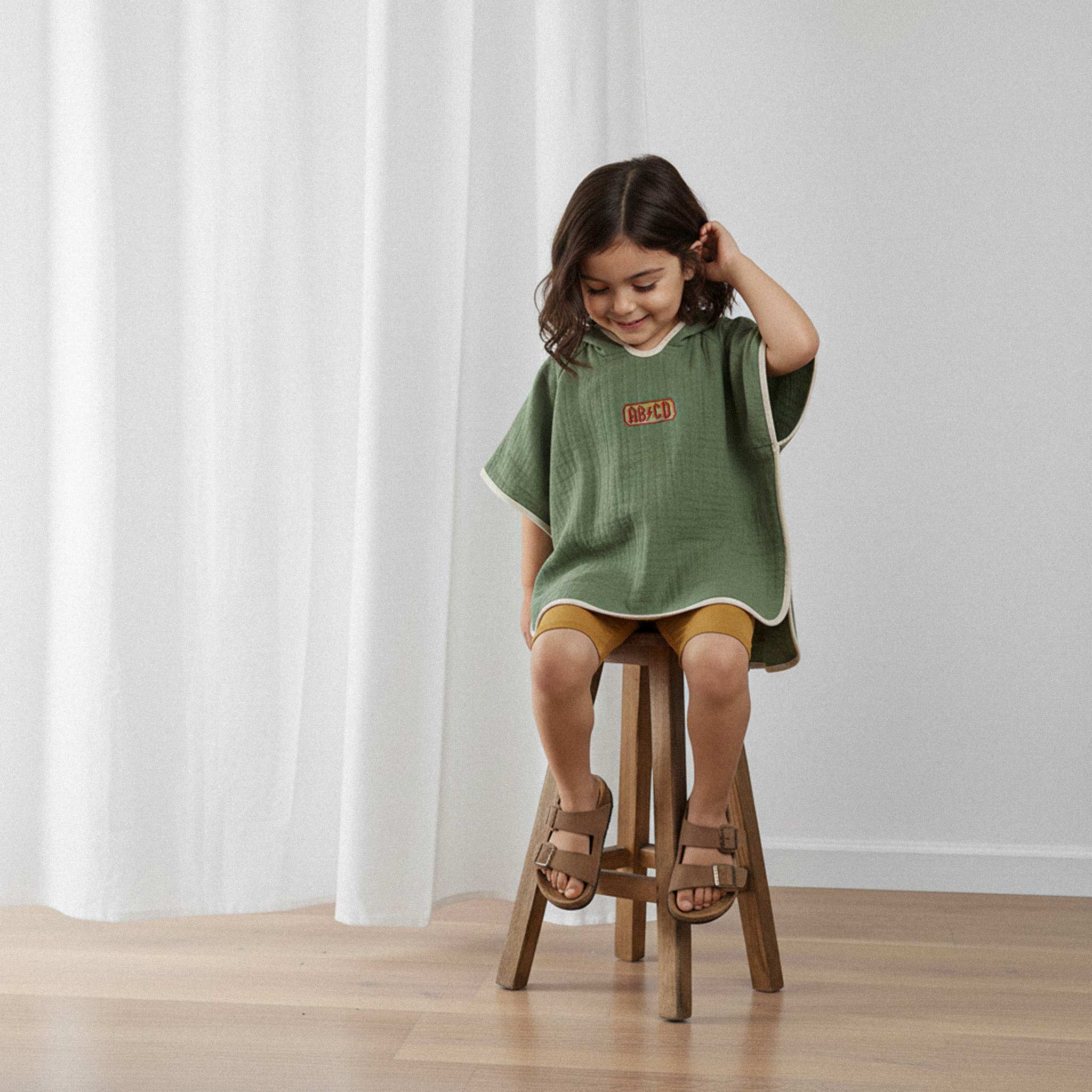 Üzerinde haki yeşili, 'AB/CD' nakışlı kapüşonlu müslin panço bulunan küçük kız çocuk, ahşap bir taburede otururken gülümseyerek pançosuna bakıyor.