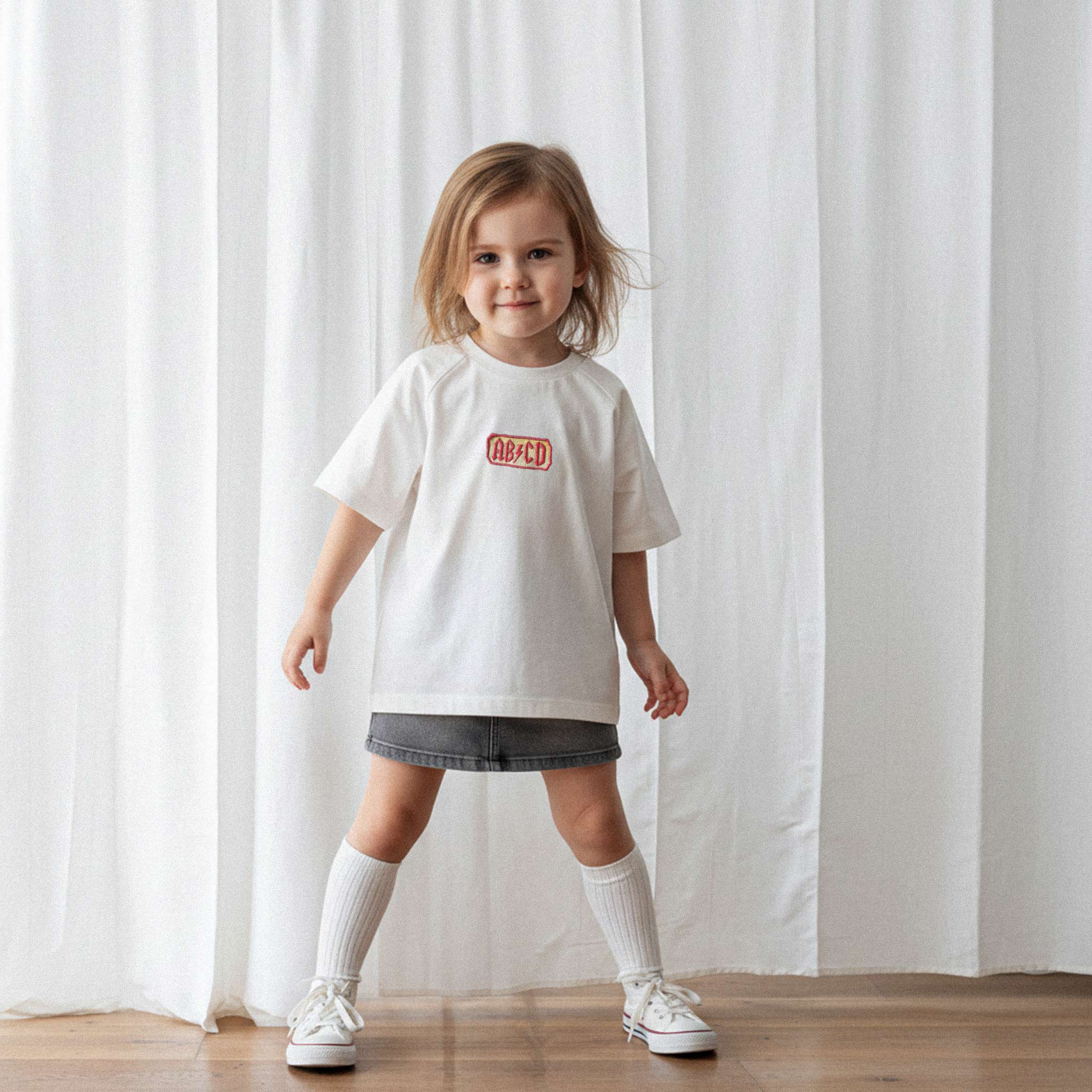 Üzerinde ekru renkli, reglan kollu, 'AB/CD' nakışlı oversize bebek tişörtü bulunan kız çocuk, kameraya doğru gülümseyerek poz veriyor.