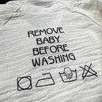 Açık bej renkli %100 pamuklu müslin çocuk kimono takımı. Üzerinde gri iplikle işlenmiş esprili "REMOVE BABY BEFORE WASHING" mesajı ve yıkama talimatı sembollerinden oluşan nakış detayı yer alır. Yüksek kalite nakış işçiliği ile üretilmiştir. Bebek ve çocukların hassas cildine uygun, nefes alan ve terletmeyen hafif yapısıyla sıcak aylar için ideal, dikkat çekici premium müslin takım.