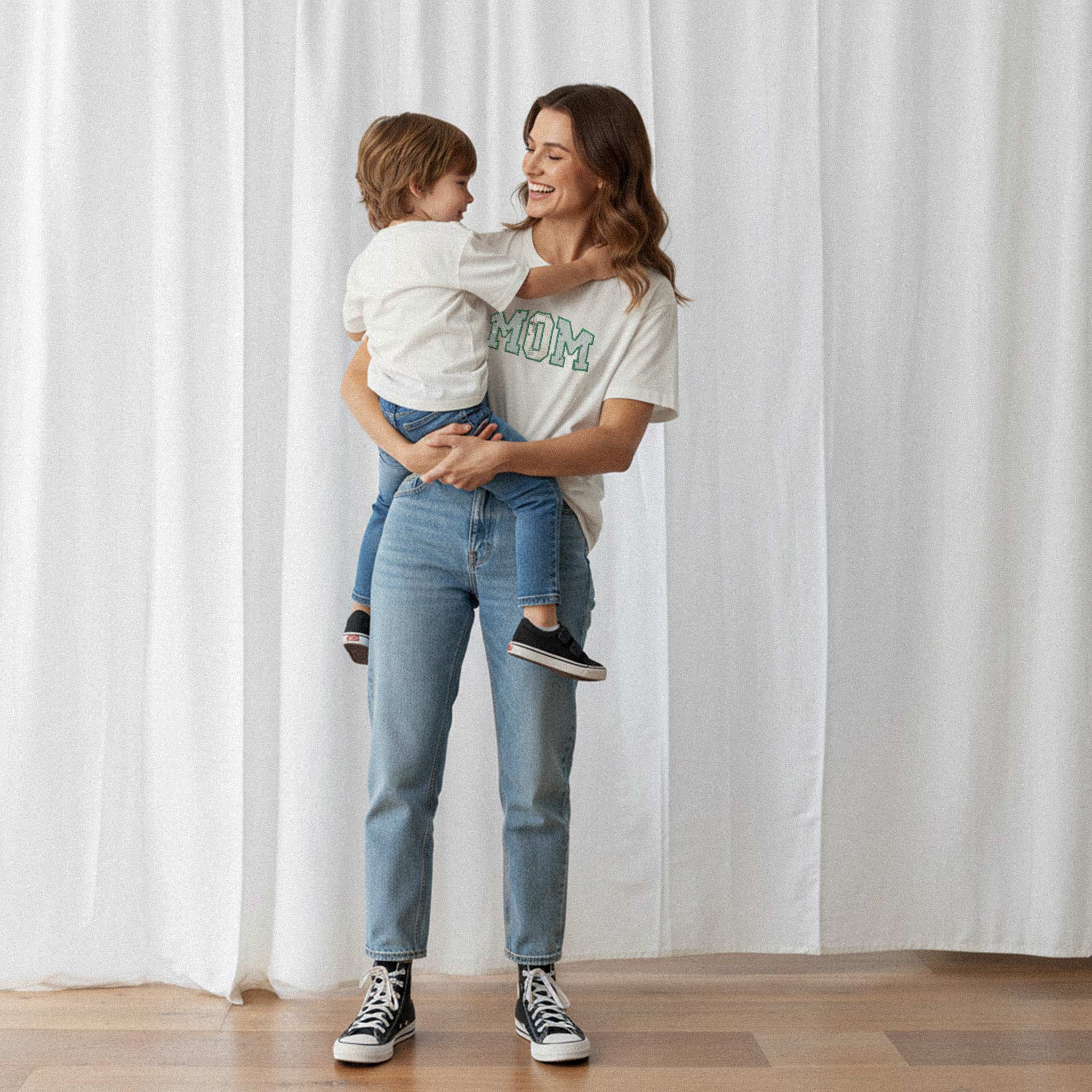 Kucağında çocuğunu tutan anne, üzerinde ekru renkli, kişiye özel 'MOM' aplikeli Bona Pera tişörtü giyiyor. Anne ve çocuğu arasındaki sevgi dolu an.