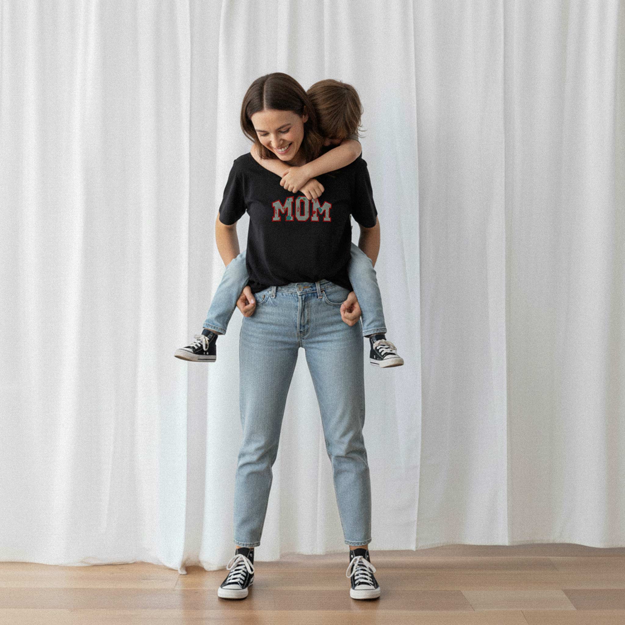 Sırtında çocuğunu taşıyan anne, üzerinde siyah renkli, kişiye özel 'MOM' aplikeli Bona Pera tişörtü giyiyor. Anne ve çocuğu arasındaki neşeli an.