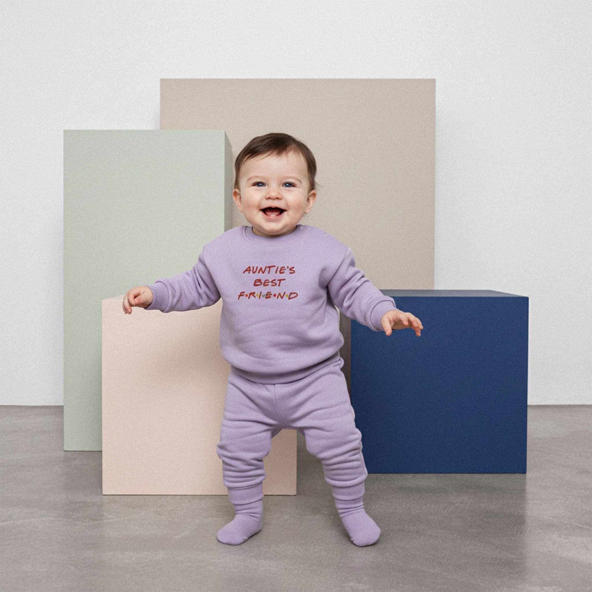 Üzerinde lila renkli, 'Auntie's Best Friend' nakışlı eşofman takımı bulunan sevimli bebek, neşeyle gülümseyerek ayakta duruyor.