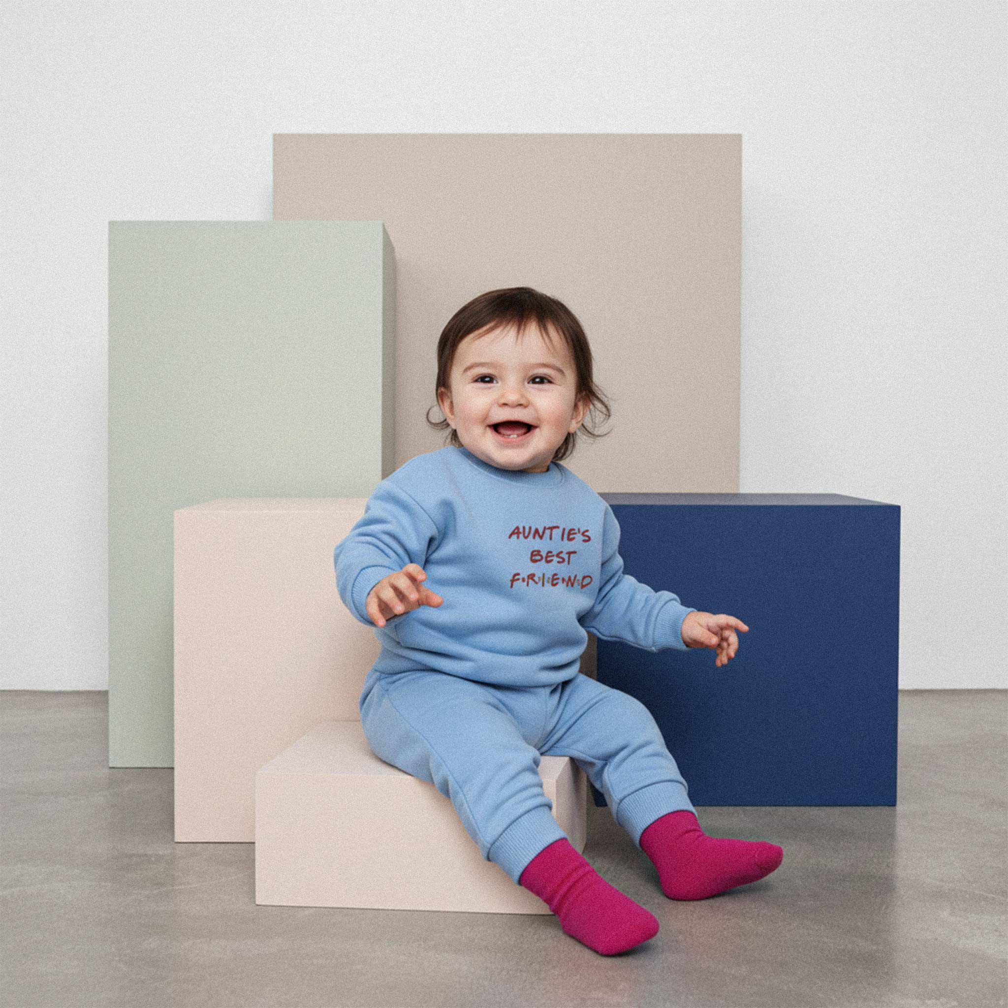 Üzerinde mavi renkli, 'Auntie's Best Friend' nakışlı eşofman takımı bulunan sevimli bebek, renkli küplerin üzerinde otururken neşeyle gülümsüyor.