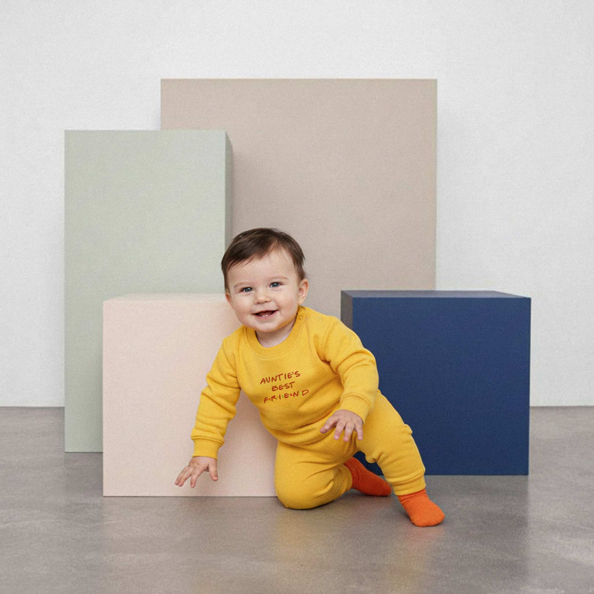 Üzerinde hardal sarısı renkli, 'Auntie's Best Friend' nakışlı eşofman takımı bulunan sevimli bebek, renkli küplerin önünde emeklerken neşeyle gülümsüyor.