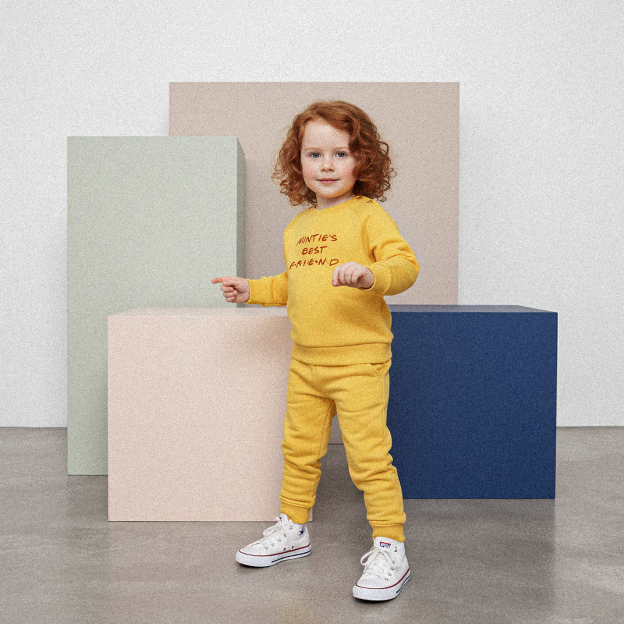 Üzerinde hardal sarısı renkli, 'Auntie's Best Friend' nakışlı eşofman takımı bulunan kızıl saçlı sevimli çocuk, renkli küplerin önünde poz veriyor.