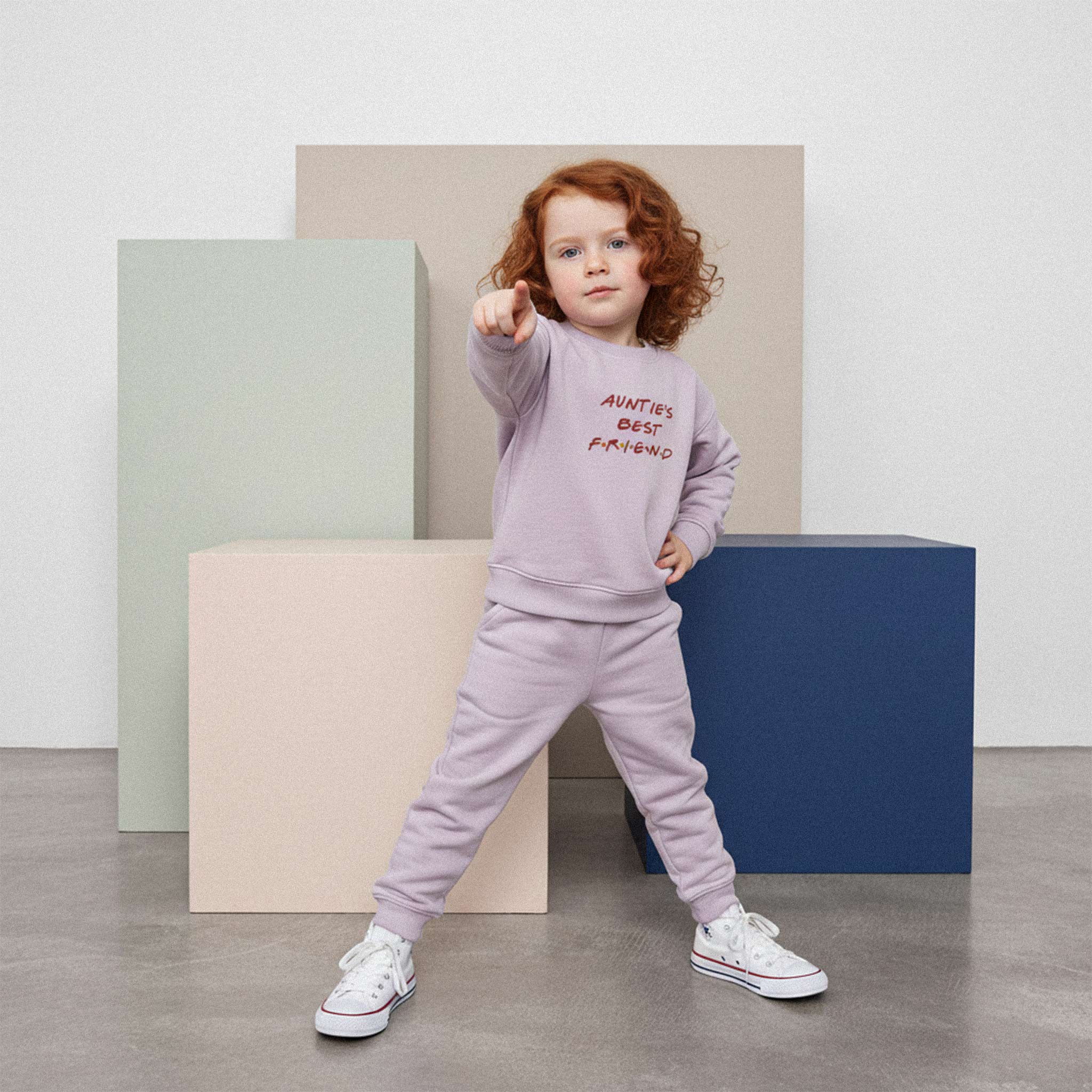 Kızıl saçlı, lila renkli 'Auntie's Best Friend' eşofman takımı giyen kız çocuk, elini beline koyarak ve kamerayı işaret ederek havalı bir poz veriyor.