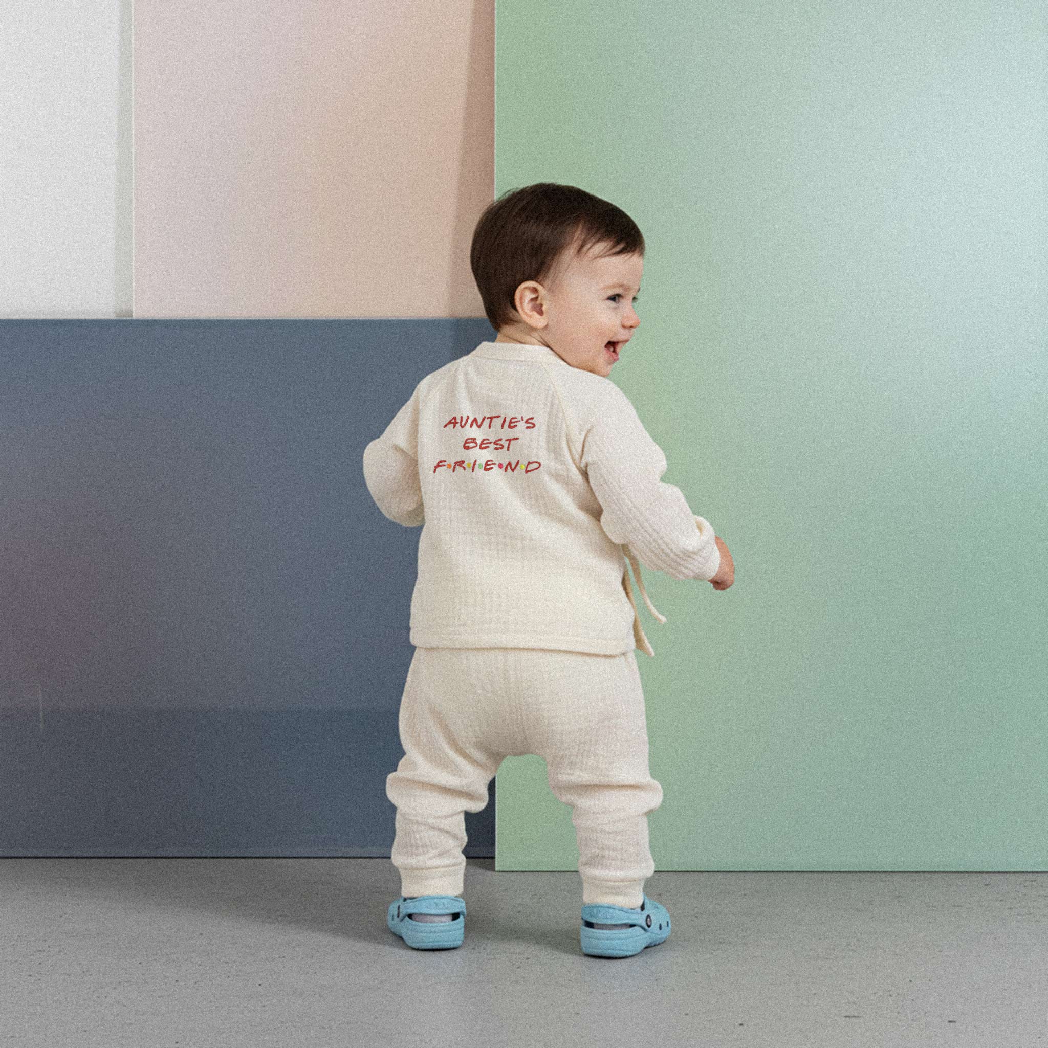 Üzerinde ekru renkli, 'Auntie's Best Friend' baskılı müslin kimono takımı bulunan sevimli bebek, renkli küplerin önünde ayakta durarak neşeyle gülümsüyor.