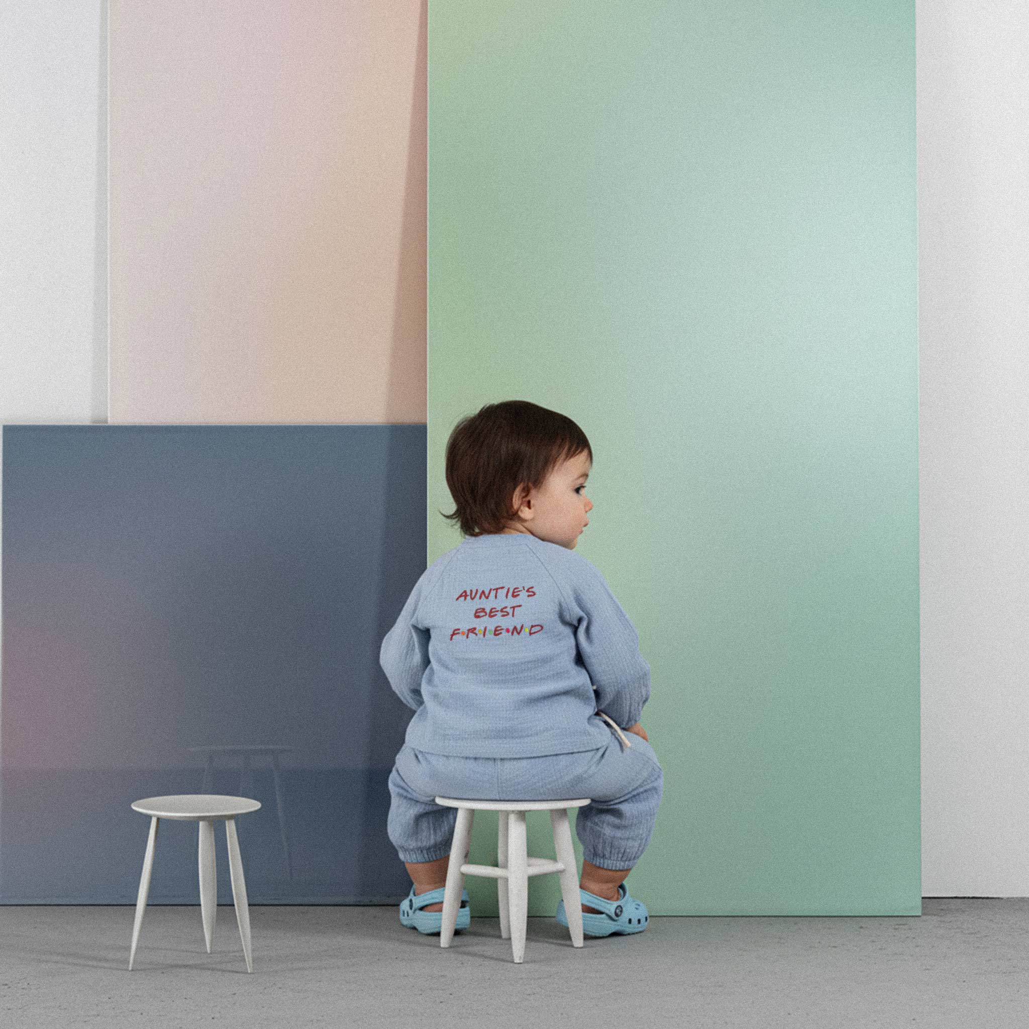 Üzerinde indigo mavisi renkli, 'Auntie's Best Friend' baskılı müslin kimono takımı bulunan sevimli bebek, küçük bir taburede otururken merakla kameraya bakıyor.