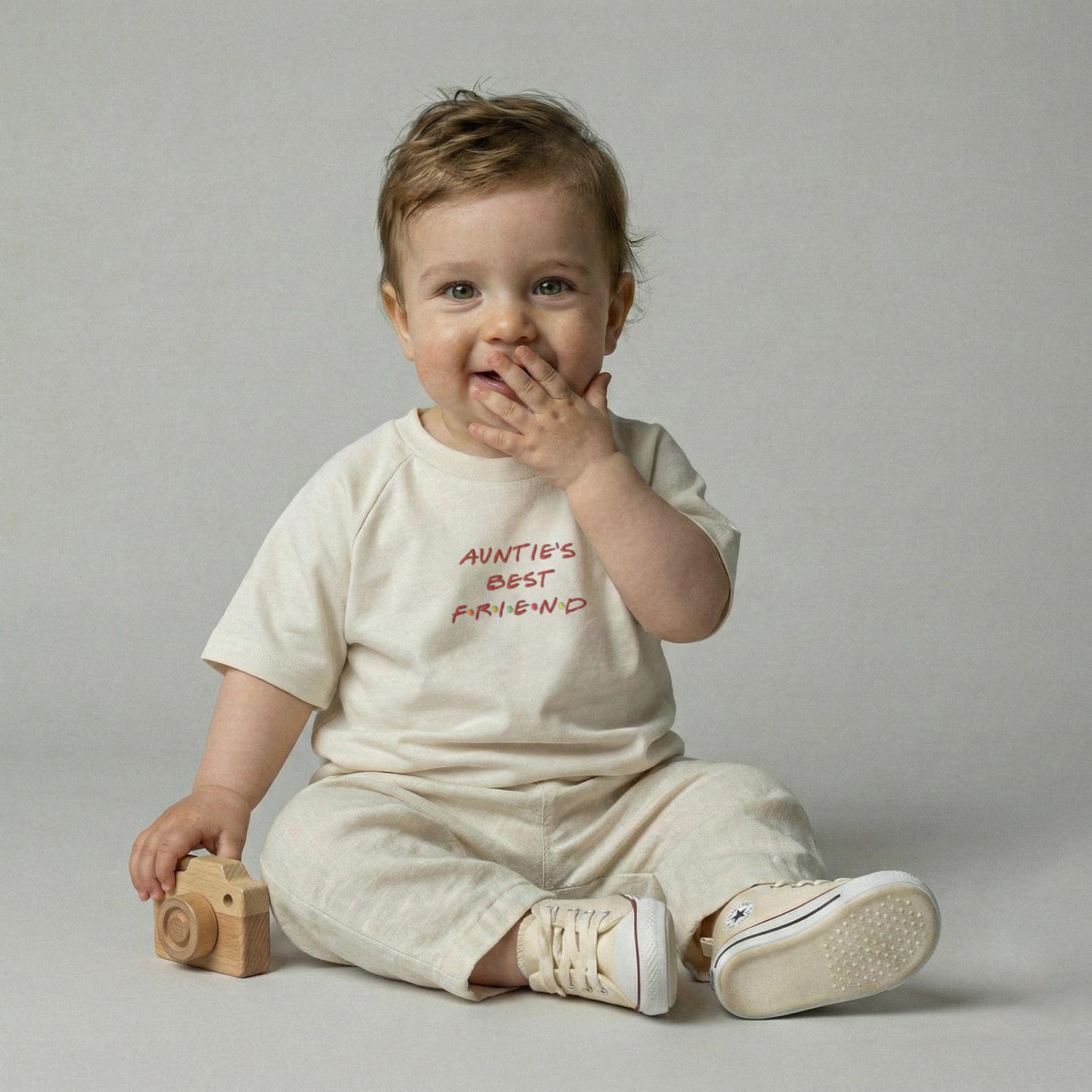 Ekru renkli, 'Auntie's Best Friend' nakışlı pamuklu tişört ve takım pantolon giyen, elinde ahşap oyuncak kamera tutarak kıkırdayan erkek bebek model.