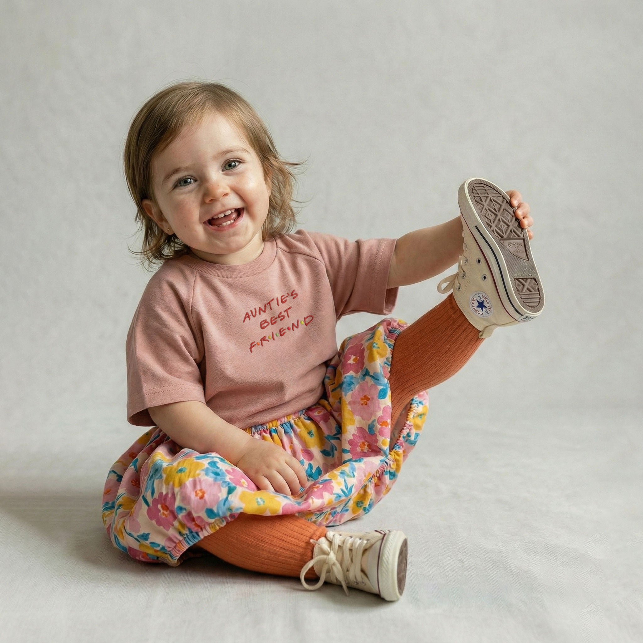 Gül kurusu renginde 'Auntie's Best Friend' nakışlı tişört ve çiçekli etek giyen, ayağındaki ayakkabıyı tutarak neşeyle gülen kız bebek model.
