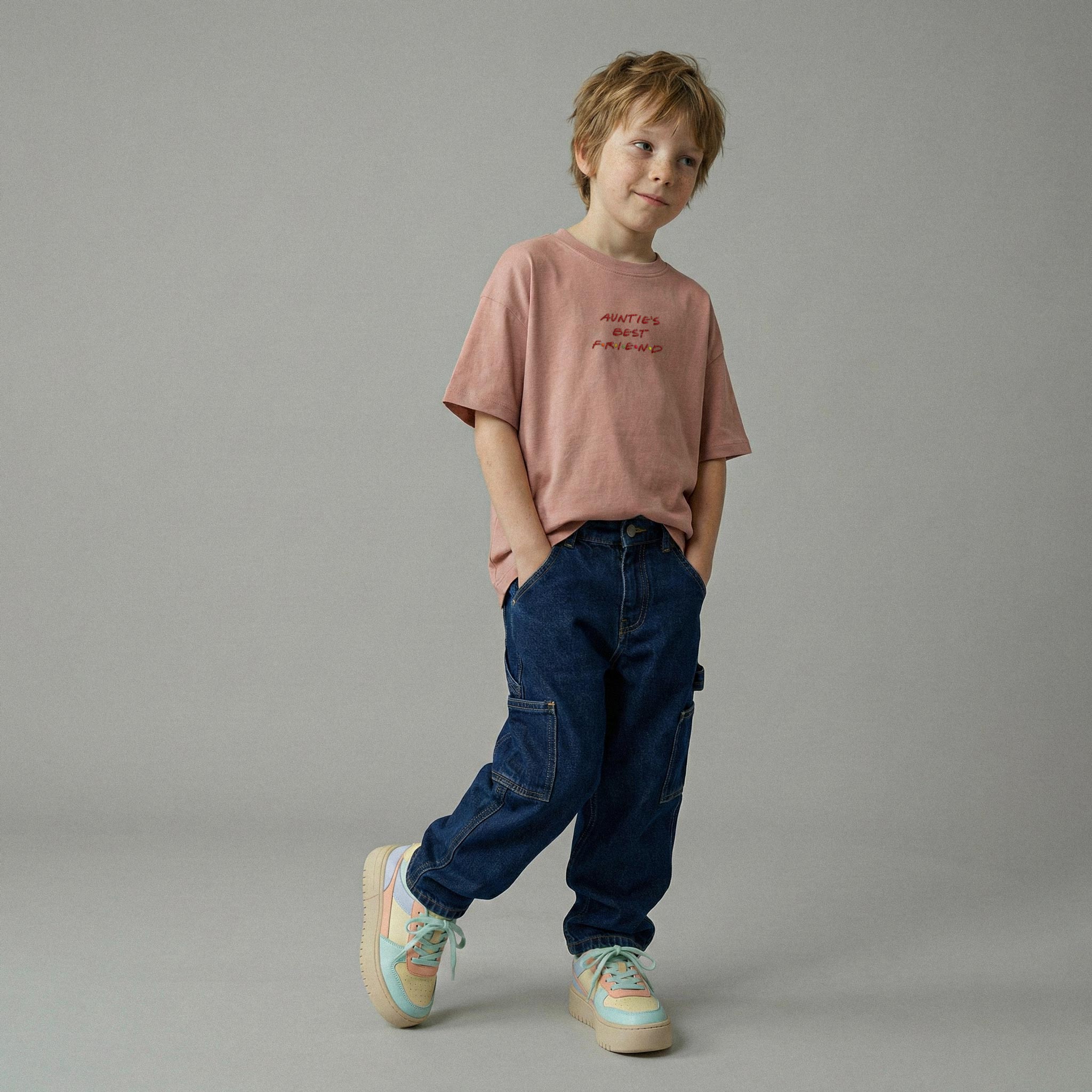 Gül kurusu renginde 'Auntie's Best Friend' nakışlı oversize tişört ve lacivert kargo jean giyerek elleri cebinde havalı bir şekilde poz veren erkek çocuk model.