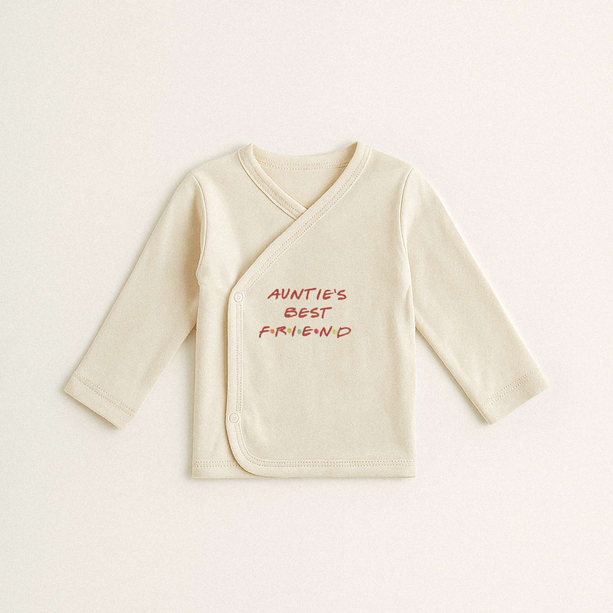 Beyaz arka plan üzerinde duran, ekru renkli, %100 pamuklu, uzun kollu kruvaze yenidoğan üst. Üzerinde Friends dizisi stilinde kırmızı harflerle "AUNTIE'S BEST FRIEND" yazısı bulunmaktadır.
