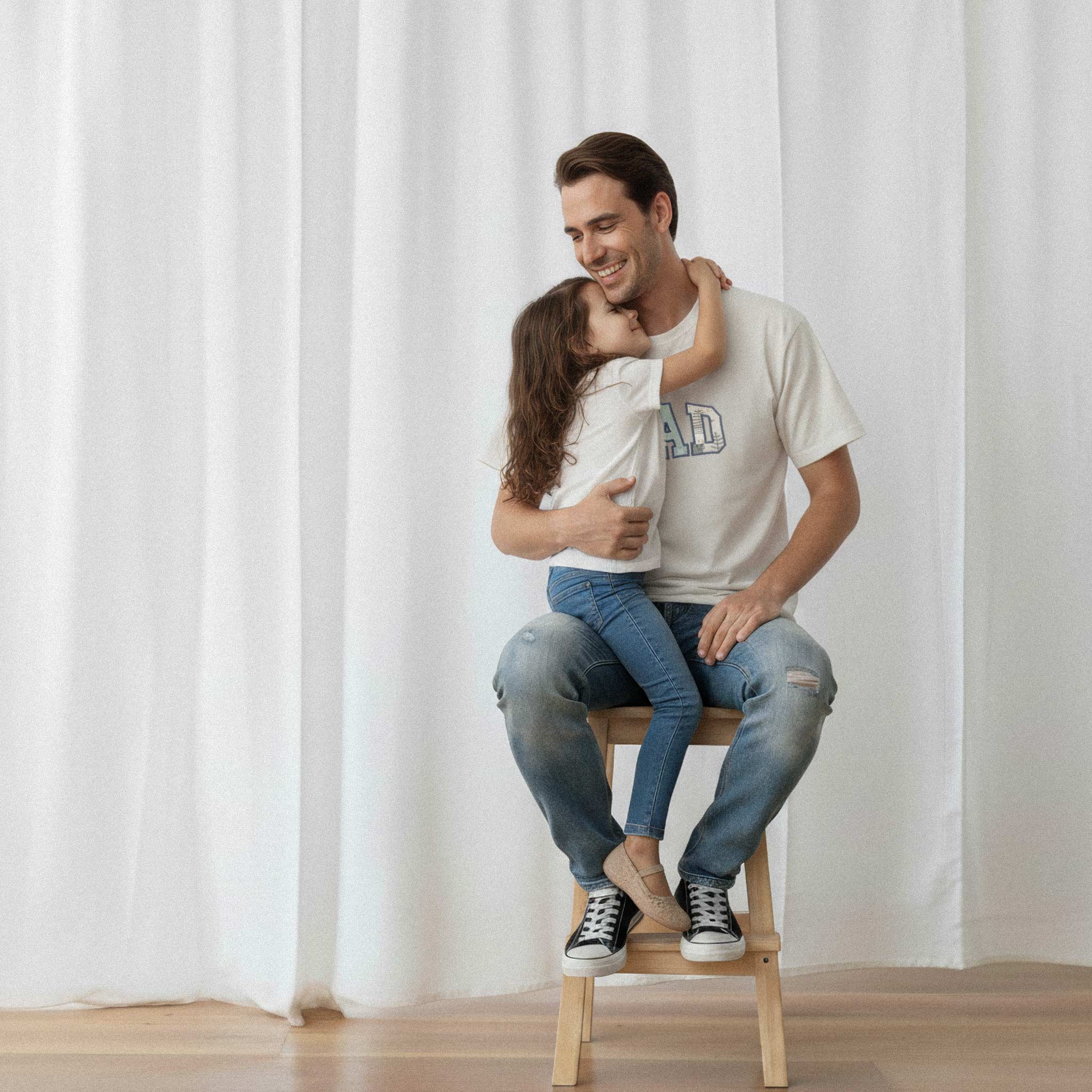 Kucağında oturan kızına sarılan baba, üzerinde ekru renkli, kişiye özel 'DAD' aplikeli Bona Pera tişörtü giyiyor. Baba ve kızı arasındaki sevgi dolu an.