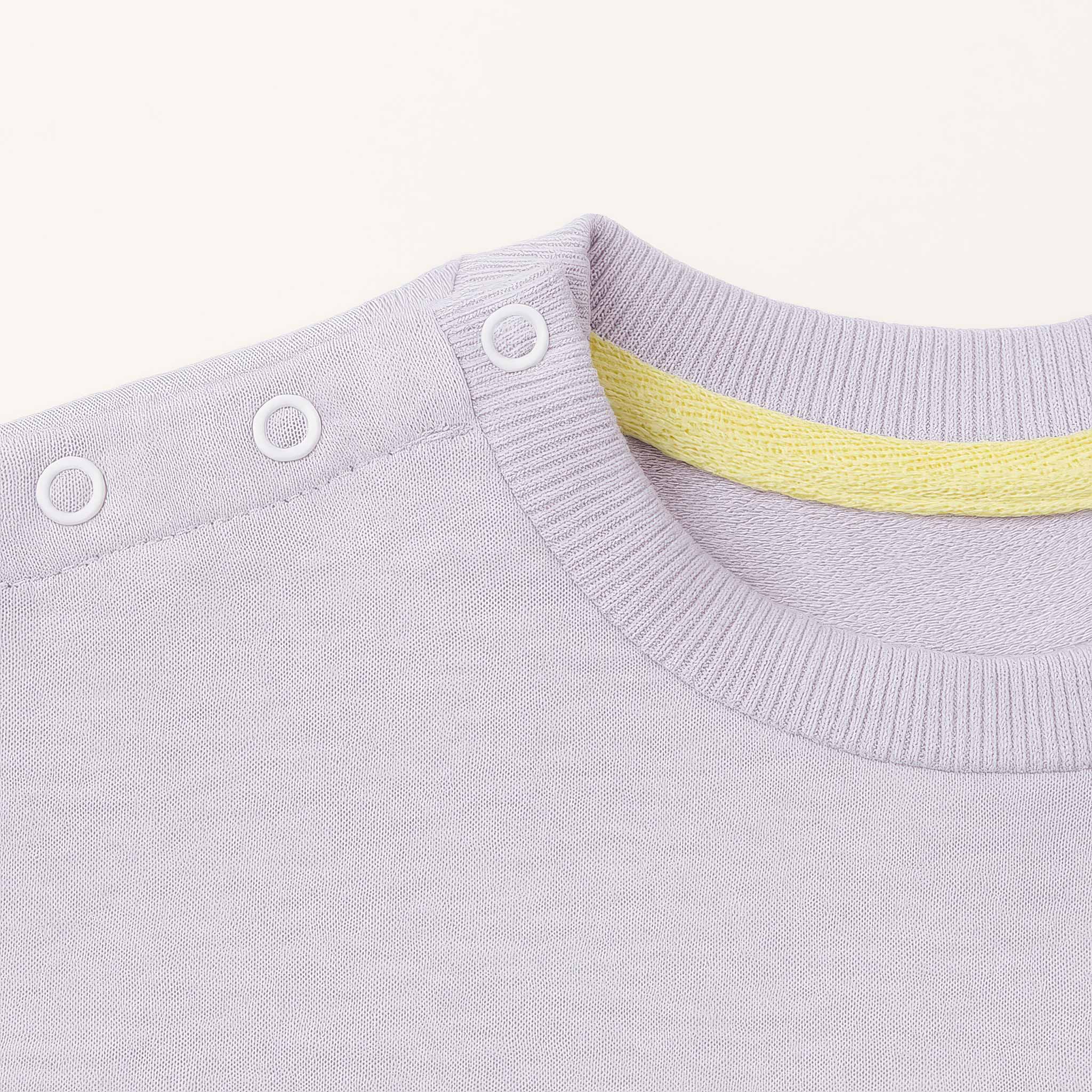 Lila renkli bebek sweatshirt'ünün omuz kısmındaki beyaz çıtçıtların ve yaka içindeki sarı biyenin yakın plan kalite detayı fotoğrafı.