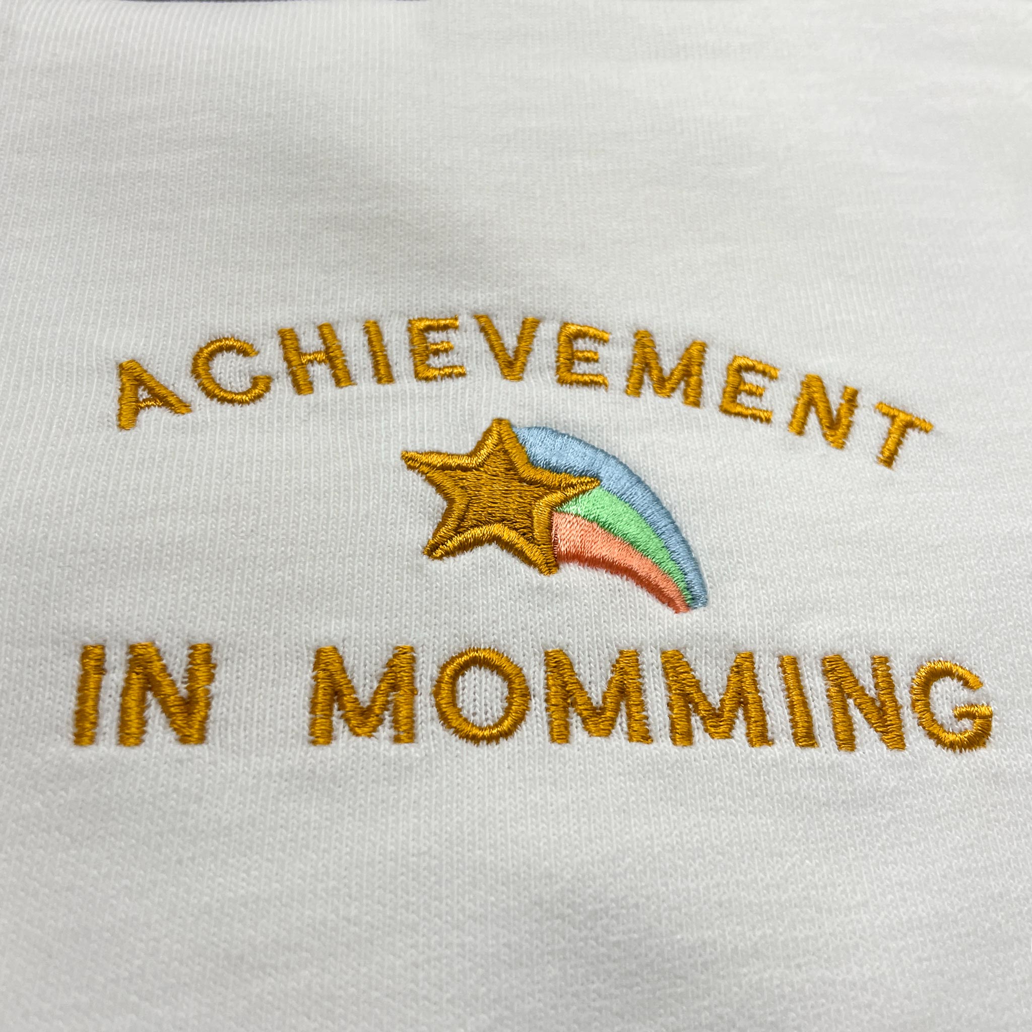 Kırık beyaz renkli %100 pamuklu premium kadın loose fit tişört üzerinde yer alan "Achievement in Momming" yazısı ve çok renkli kayan yıldız motifli nakış detay görünümü. Nakış işçiliği ile özenle işlenmiştir. Oeko-Tex sertifikalı heavyweight kumaşı sayesinde tok bir duruş sergiler, cilde nefes aldırır ve terletmez. Anneliği eğlenceli bir dille kutlayan, gün boyu konfor sağlayan dayanıklı ve dökümlü günlük giyim tasarımı.
