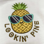 Beyaz dokulu pamuklu kumaş üzerine işlenmiş "LOOKIN' PINE" ananas nakışının makro (yakın çekim) detayı. Görselde mavi çerçeveli güneş gözlüğü takan, gülümseyen ananas figürünün ve etrafındaki kiremit/gold tonlarındaki yazıların yoğun, sıkı dikiş dokusu net bir şekilde görülmektedir. İpliklerin canlılığı ve birinci sınıf Alman nakış ipliklerinin kalitesini gözler önüne seren, eğlenceli ve premium tasarımın yüksek çözünürlüklü detay karesi.