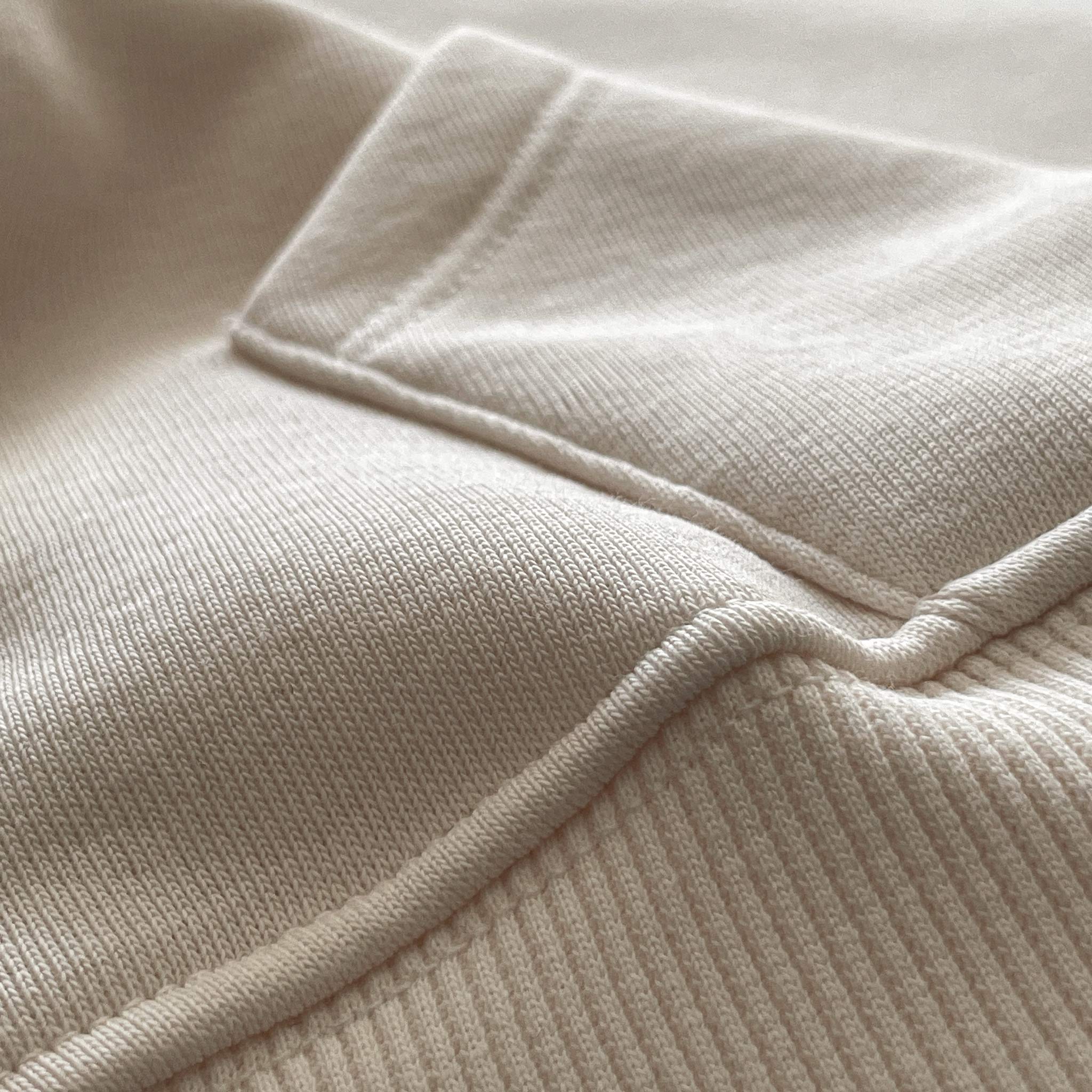 Yetişkinler için kapüşonlu beyaz hoodie modelinin minimalist cep dikişini ve kumaş dokusunu gösteren yakın çekim. Kaliteli tok kumaşın örgü dokusu ve temiz işçilikle hazırlanmış cep kenar detayı. Şıklık ve konfor sunan cep tasarımının temel dikiş izi.