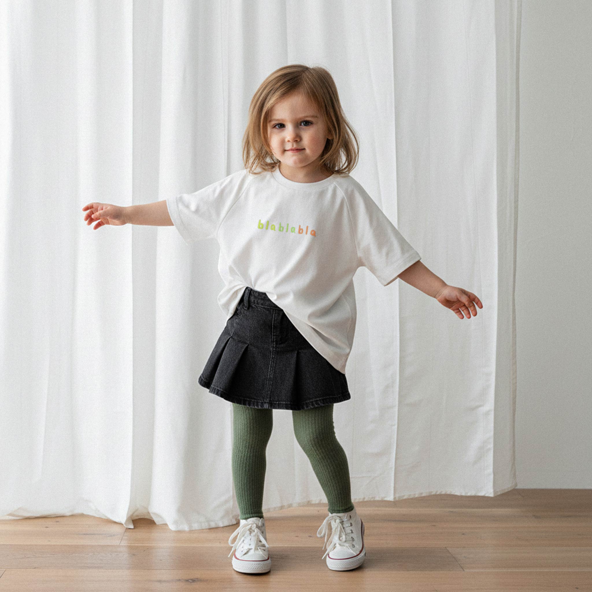 Üzerinde ekru renkli, reglan kollu, 'blablabla' nakışlı oversize bebek tişörtü bulunan küçük kız çocuk, kollarını iki yana açarak neşeyle poz veriyor.