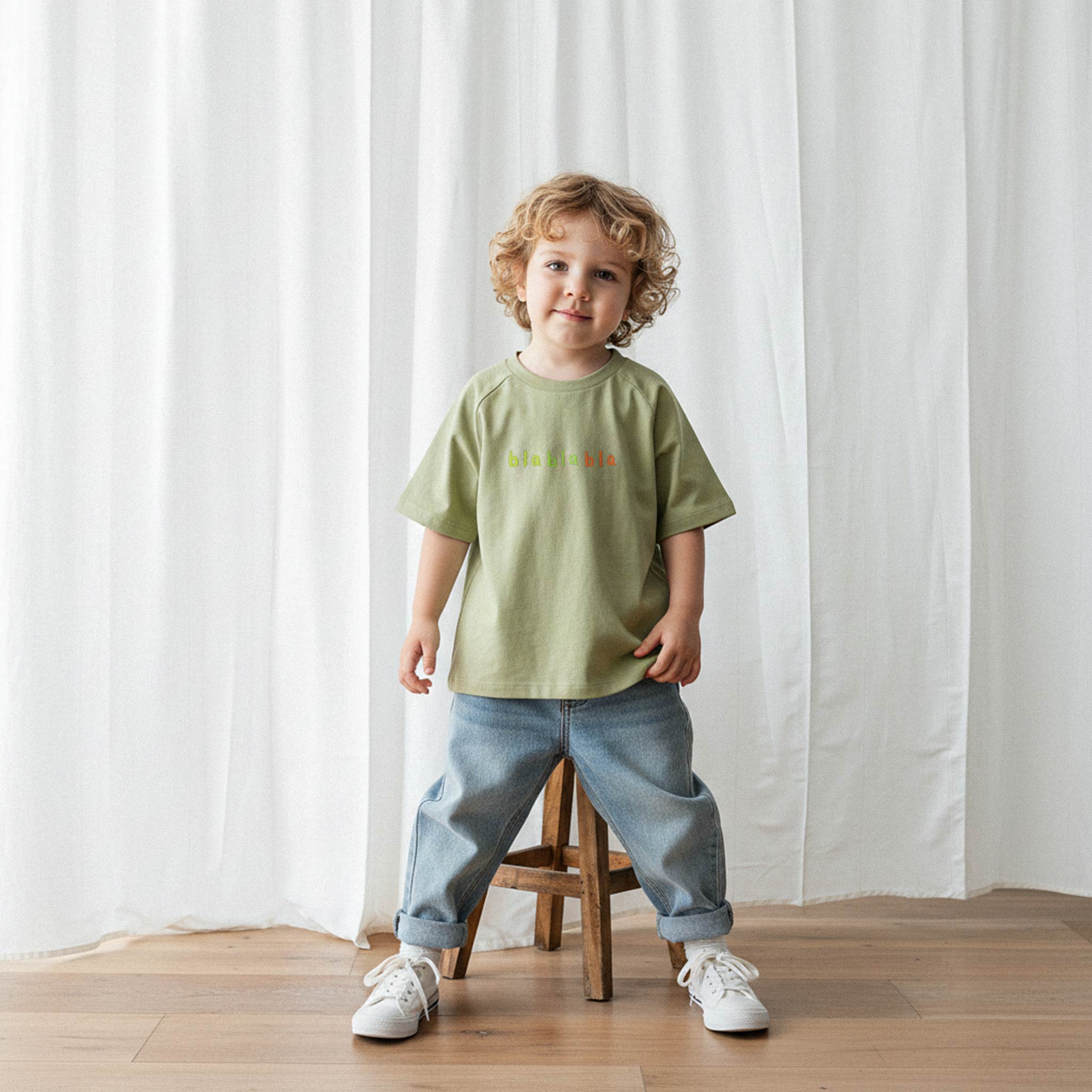 Üzerinde haki yeşili, reglan kollu, 'blablabla' nakışlı oversize bebek tişörtü bulunan kıvırcık saçlı küçük erkek çocuk, sevimli bir şekilde poz veriyor.