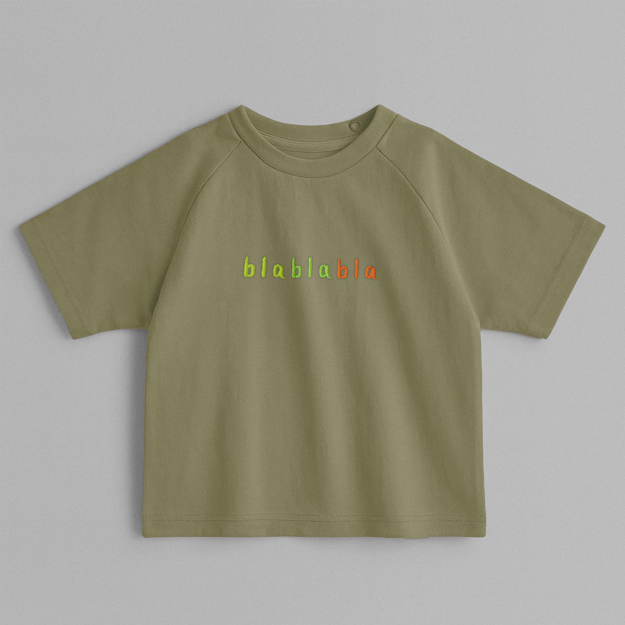 Haki yeşili, reglan kollu, omuzdan çıtçıtlı bebek tişörtü. Göğüs kısmında renkli ipliklerle "blablabla" nakışı bulunmaktadır.