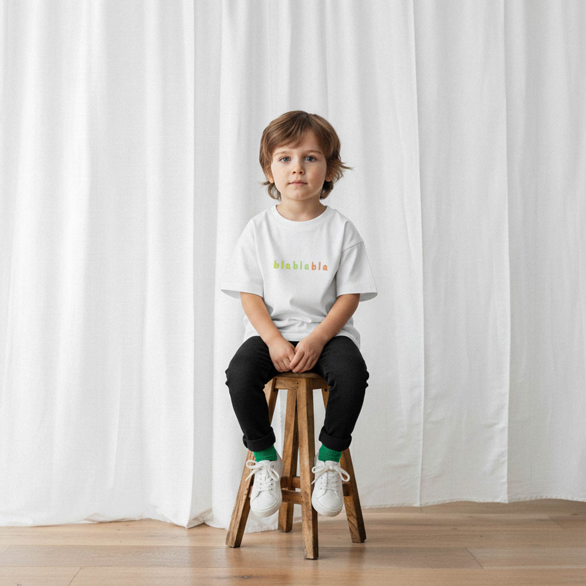 Siyah pantolon ve ekru renkli, 'blablabla' nakışlı oversize tişört giyen erkek çocuk, ahşap bir taburede oturarak kameraya bakıyor.