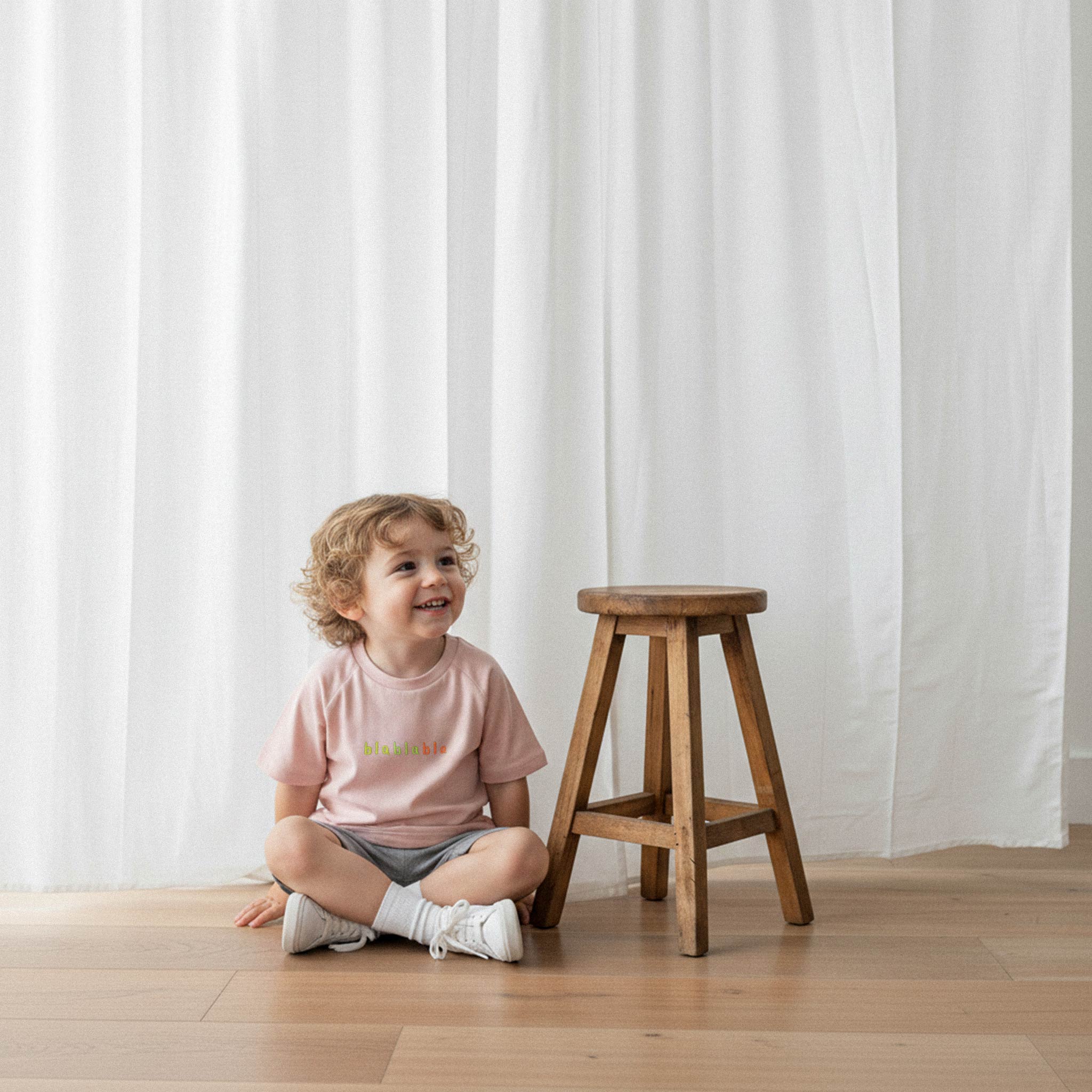 Gri şort ve gül kurusu, 'blablabla' nakışlı oversize tişört giyen kıvırcık saçlı küçük erkek çocuk, yerde otururken gülümseyerek yana bakıyor.