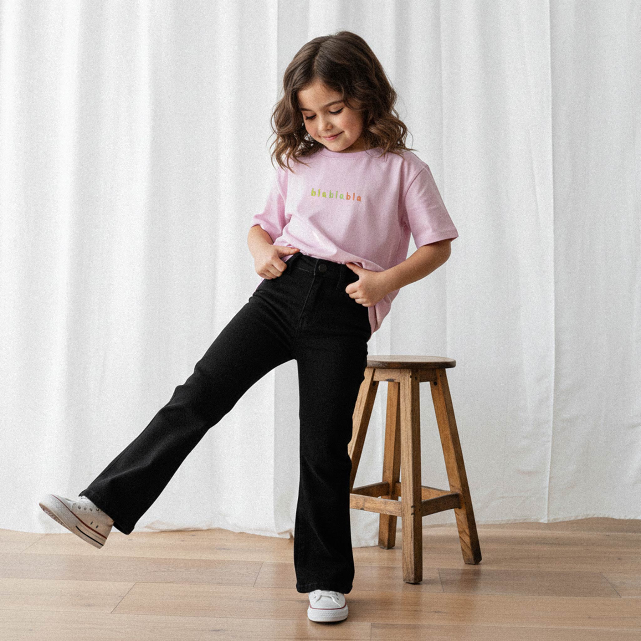 Siyah İspanyol paça pantolon ve gül kurusu, 'blablabla' nakışlı oversize tişört giyen kız çocuk, tişörtünü çekiştirerek eğlenceli bir poz veriyor.