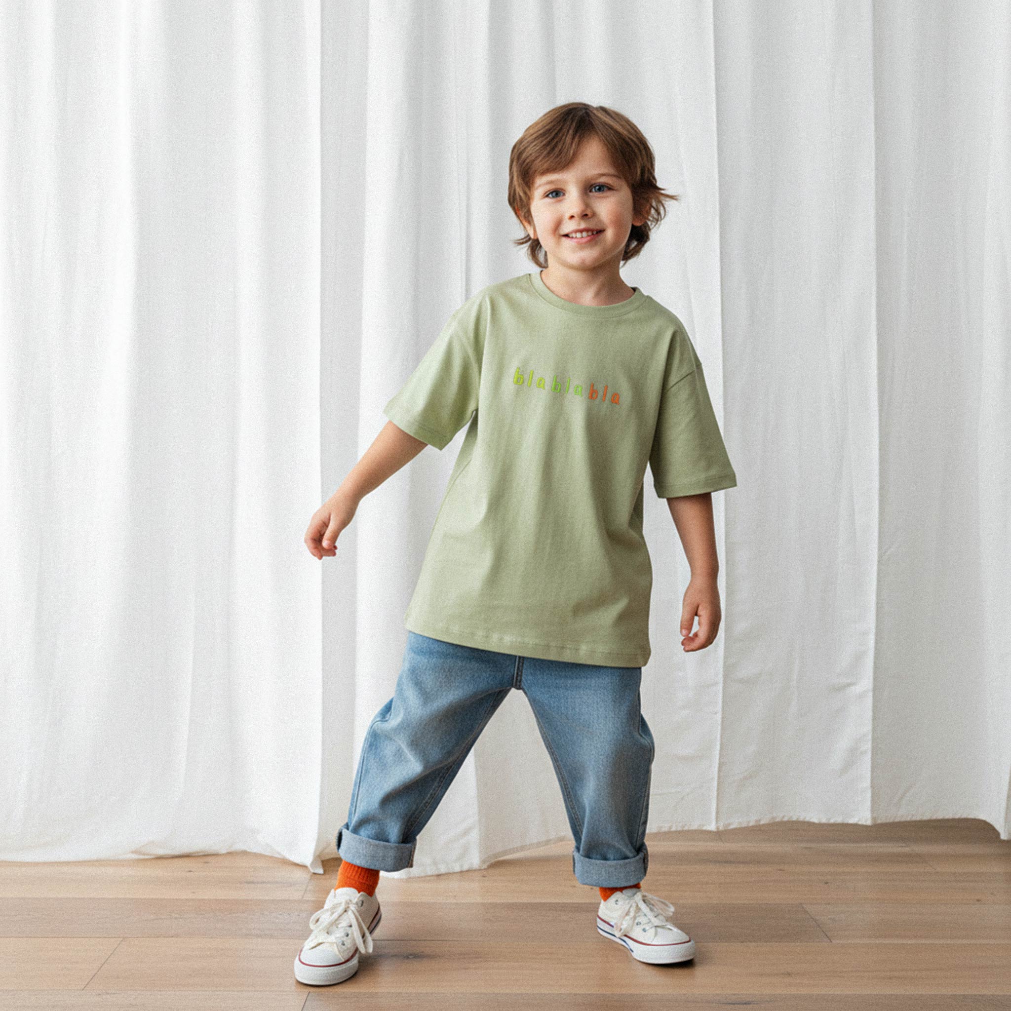 Bol paça jean pantolon ve haki yeşili, 'blablabla' nakışlı oversize tişört giyen erkek çocuk, gülümseyerek kameraya doğru yürüyor.