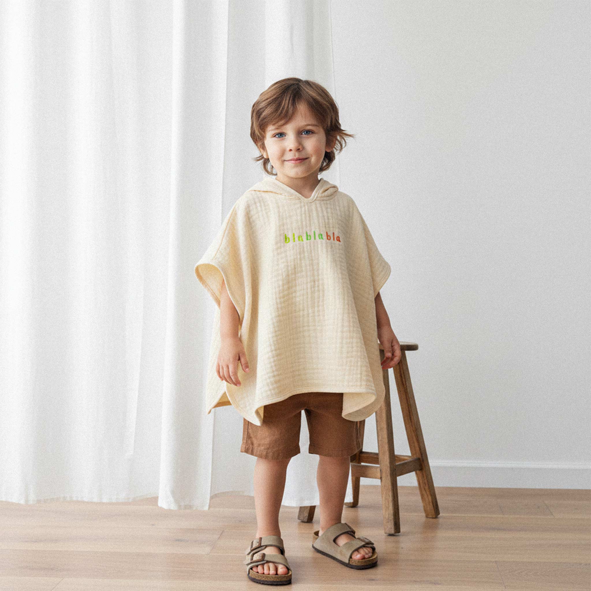 Kahverengi şort ve sandalet giyen erkek çocuk, üzerinde ekru renkli, 'blablabla' nakışlı kapüşonlu müslin panço ile gülümseyerek poz veriyor.