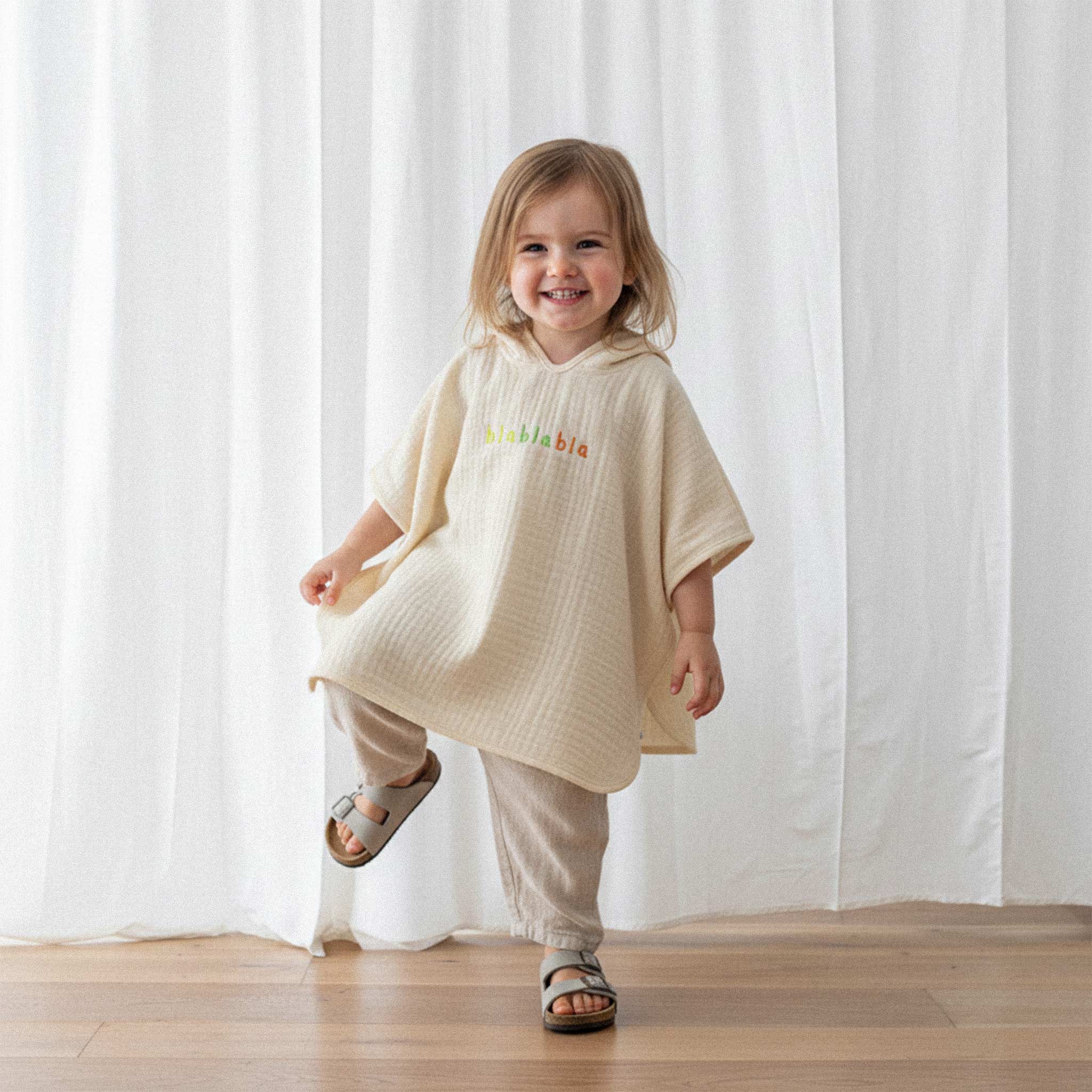 Üzerinde ekru renkli, 'blablabla' nakışlı kapüşonlu müslin panço bulunan küçük kız çocuk, tek ayağını kaldırarak neşeyle gülümsüyor.