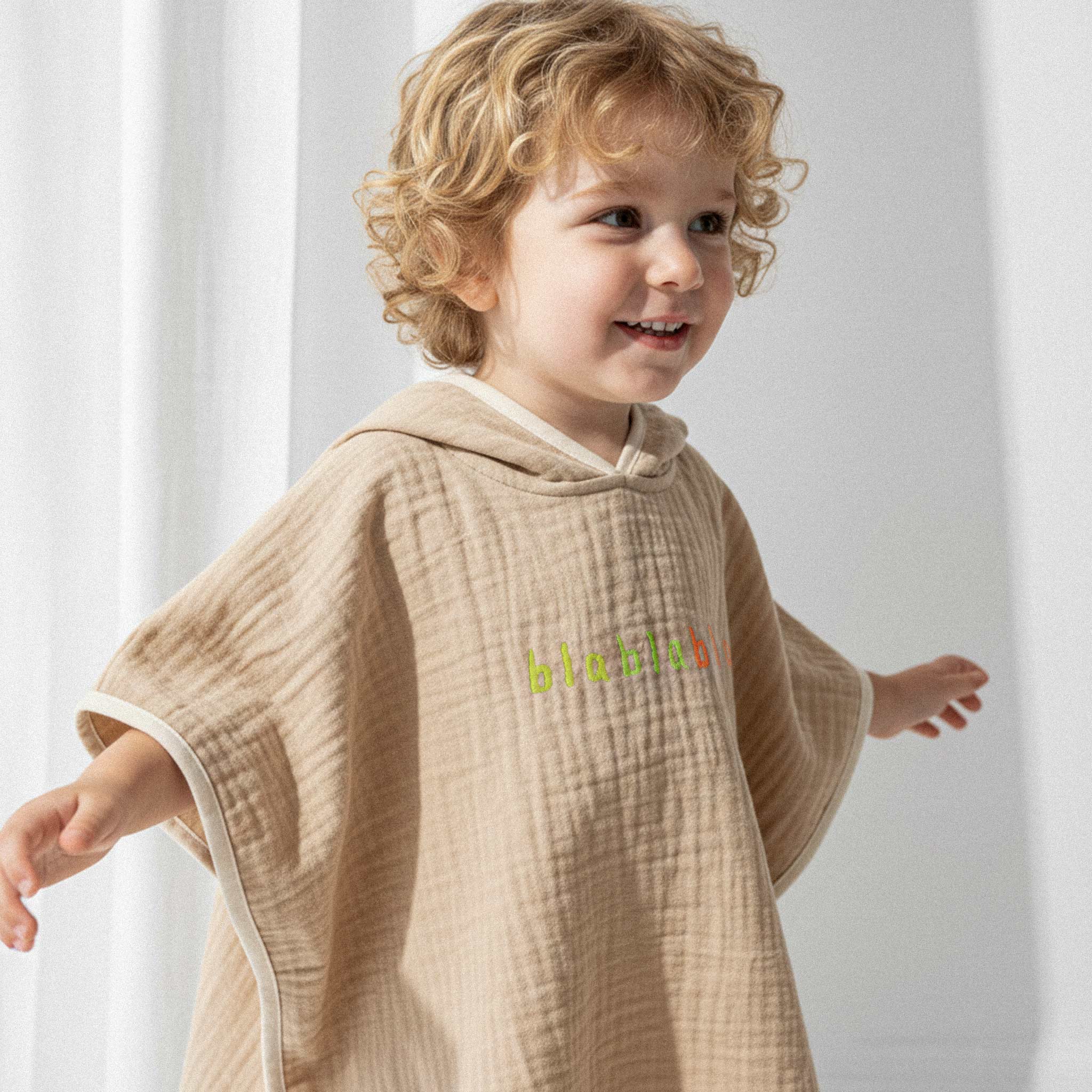 Üzerinde somon rengi, 'blablabla' nakışlı kapüşonlu müslin panço bulunan kıvırcık saçlı küçük erkek çocuk, kollarını açarak neşeyle gülümsüyor.