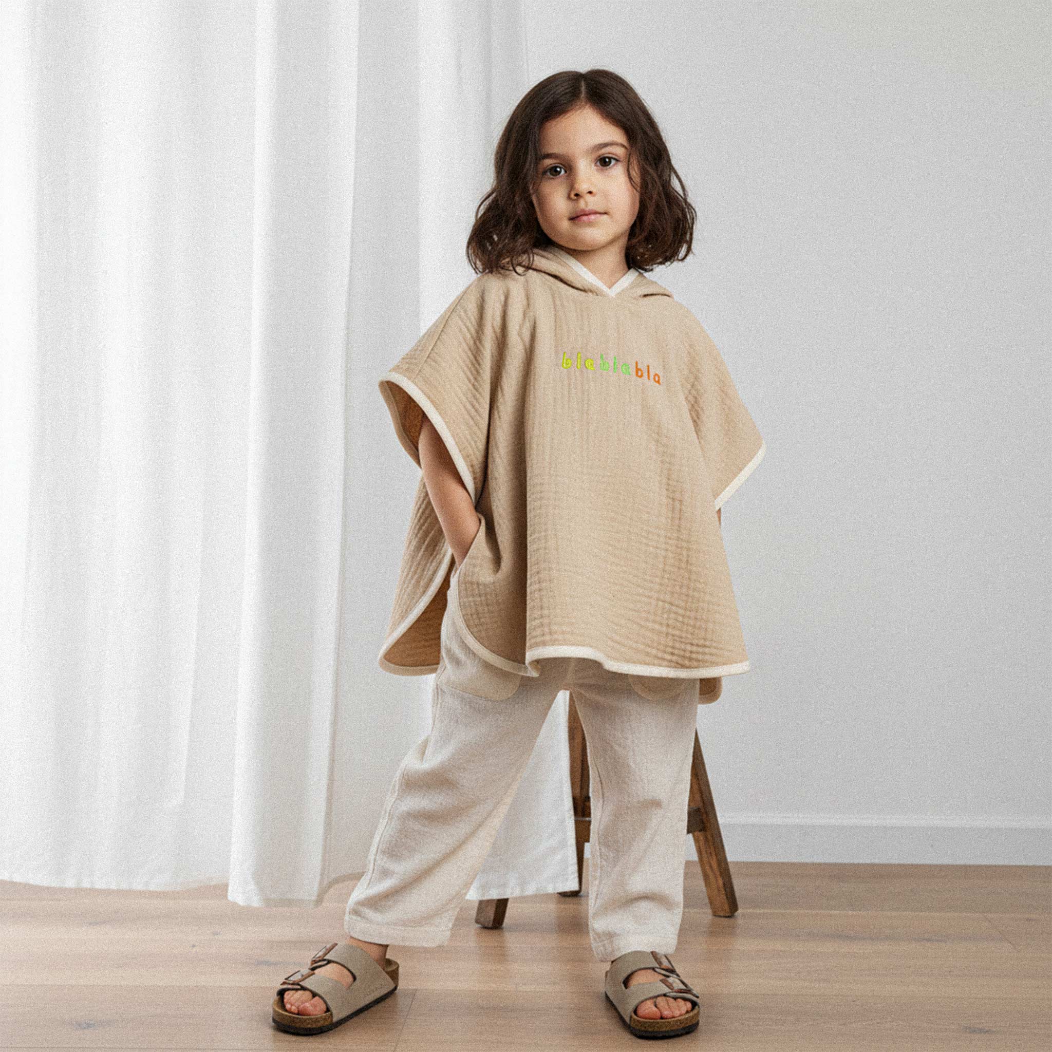 Keten pantolon giyen kız çocuk, üzerinde somon rengi, 'blablabla' nakışlı kapüşonlu müslin panço ile elleri ceplerinde havalı bir poz veriyor.