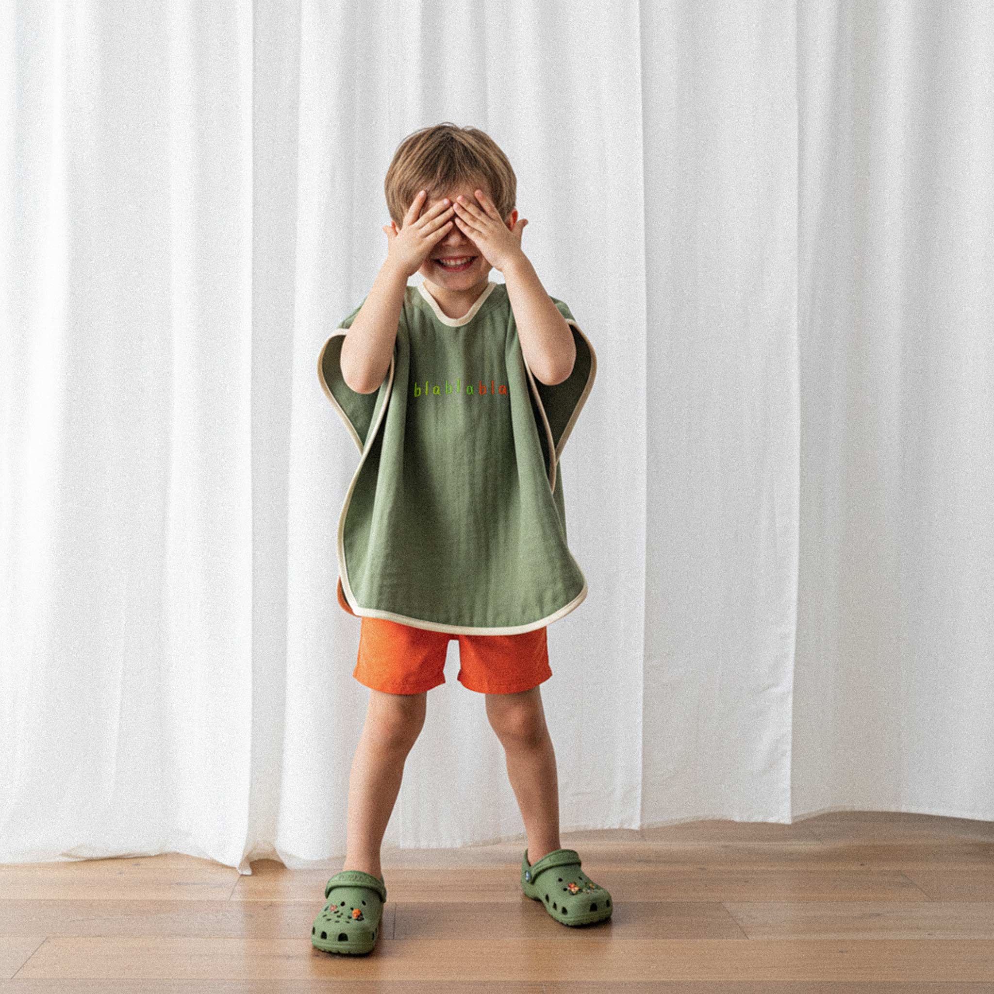 Turuncu şort ve haki yeşili, 'blablabla' nakışlı kapüşonlu müslin panço giyen erkek çocuk, gözlerini kapatarak eğlenceli bir poz veriyor.