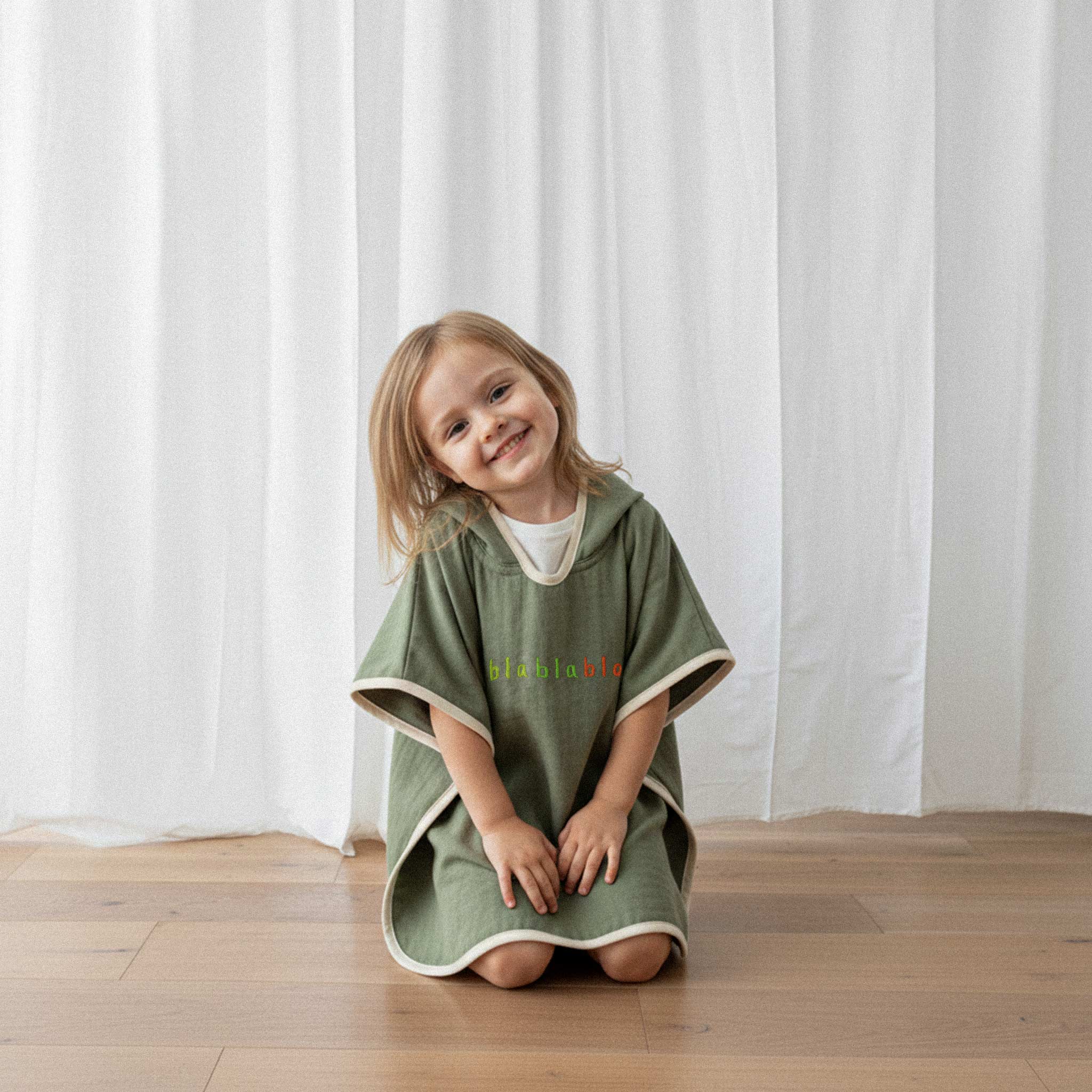 Üzerinde haki yeşili renkli, 'blablabla' nakışlı kapüşonlu müslin panço bulunan küçük kız çocuk, yerde çömelmiş gülümseyerek poz veriyor.