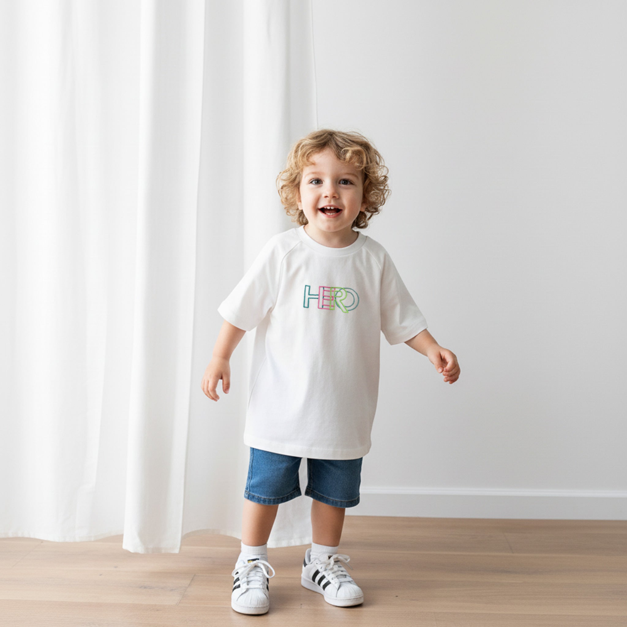 Üzerinde ekru renkli, reglan kollu, 'HERO' nakışlı oversize bebek tişörtü bulunan kıvırcık saçlı küçük erkek çocuk, neşeyle gülümseyerek yürüyor.
