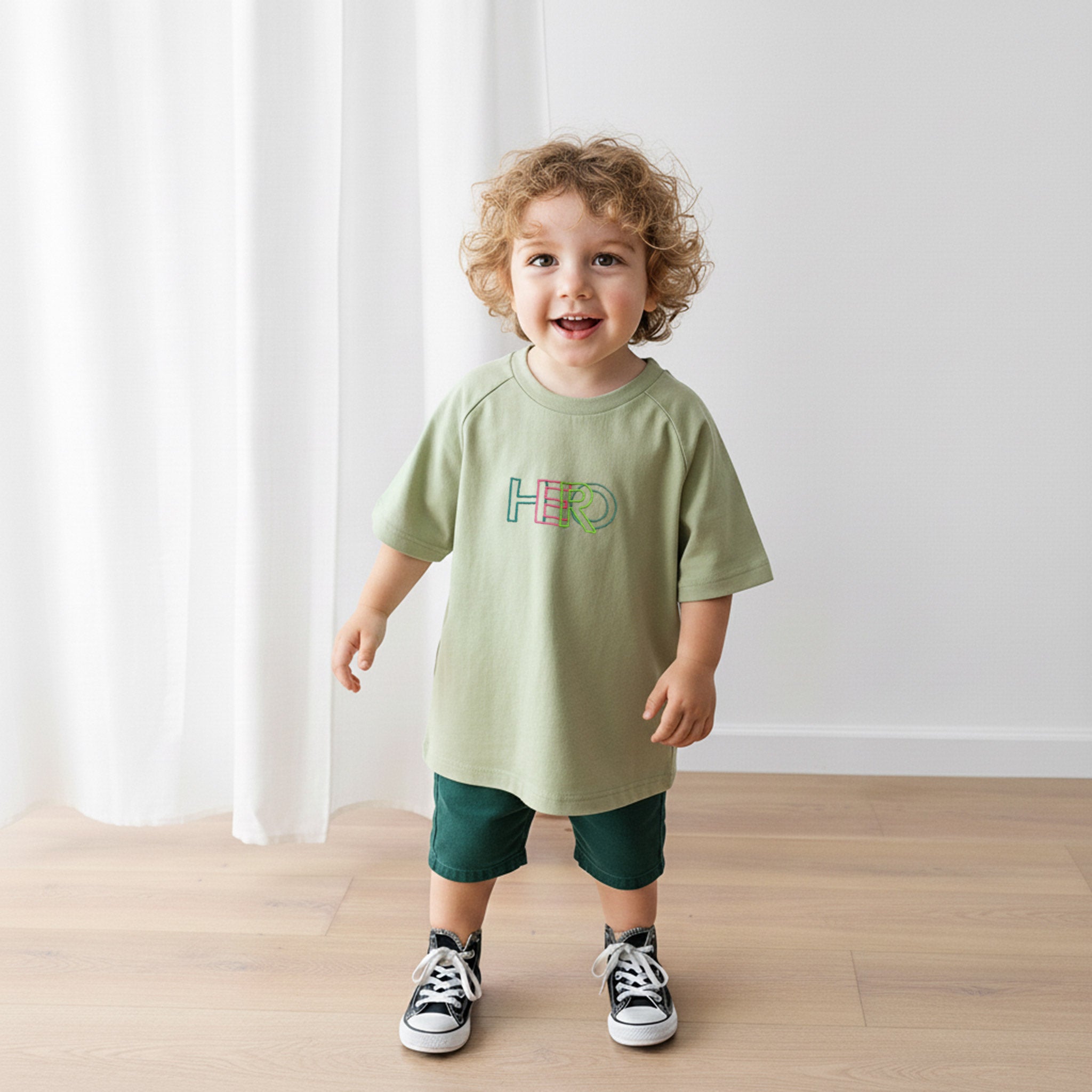 Üzerinde haki yeşili, reglan kollu, 'HERO' nakışlı oversize bebek tişörtü bulunan kıvırcık saçlı küçük erkek çocuk, neşeyle gülümseyerek yürüyor.