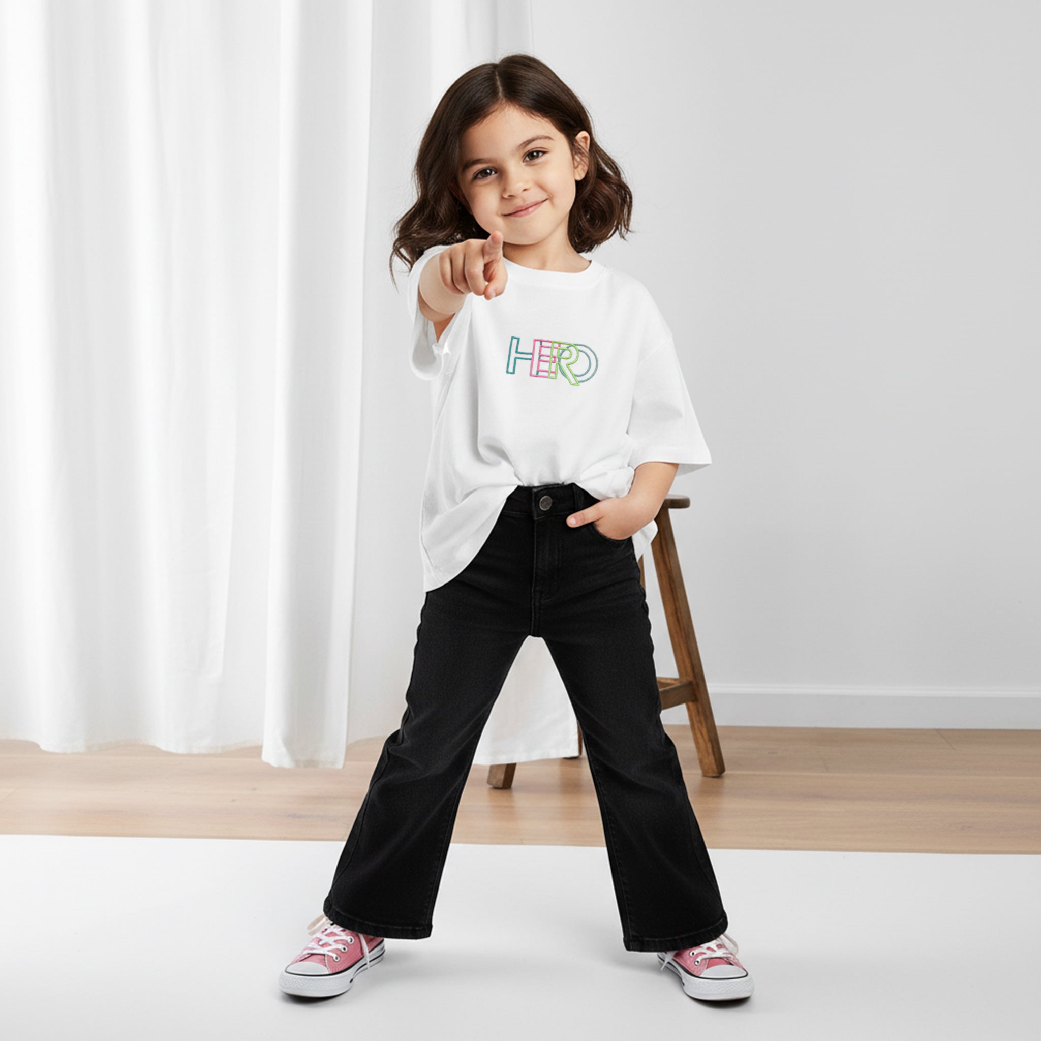 Siyah İspanyol paça pantolon ve ekru renkli, 'HERO' nakışlı oversize tişört giyen kız çocuk, gülümseyerek kamerayı işaret ediyor.