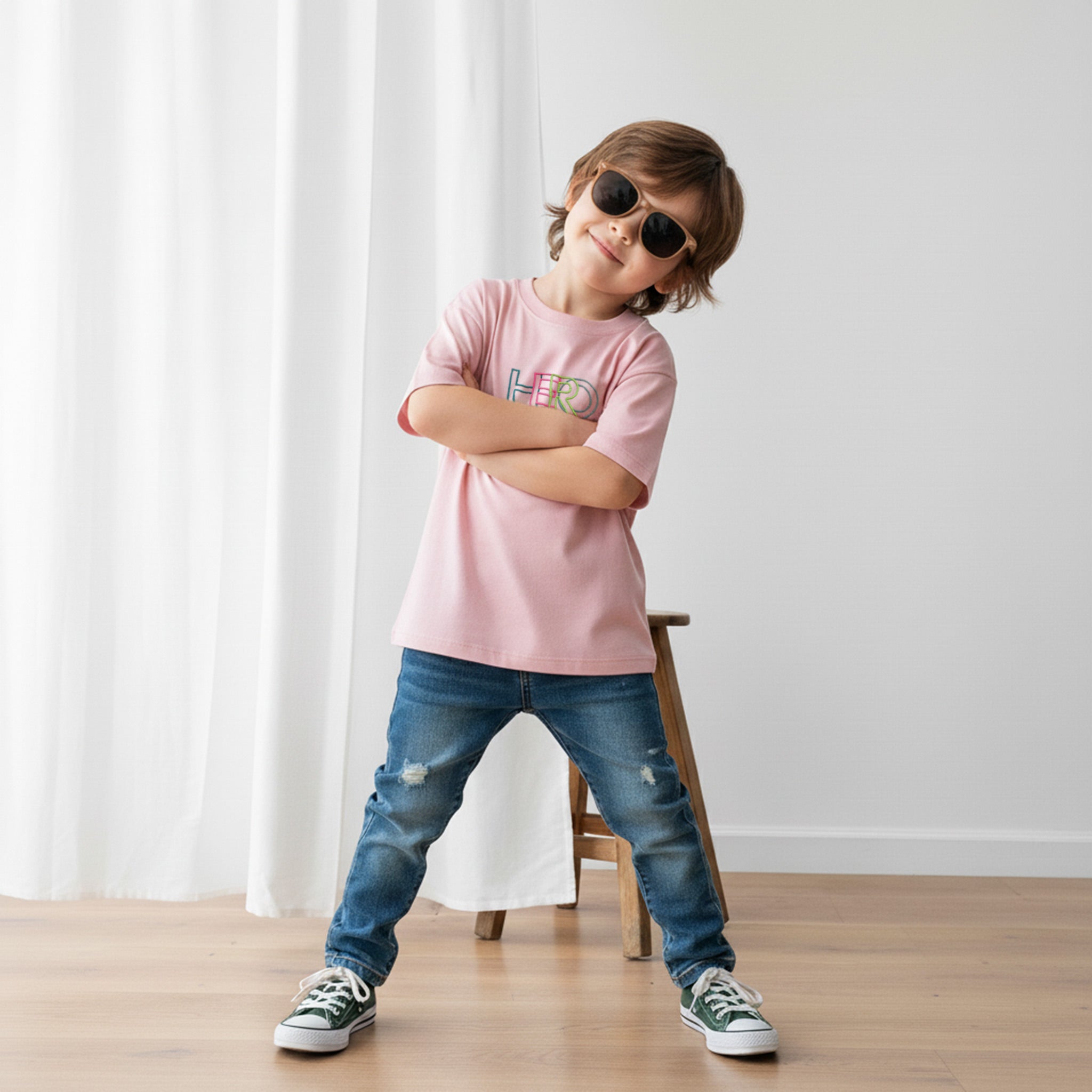 Güneş gözlüğü takan havalı erkek çocuk, üzerinde gül kurusu renkli, 'HERO' nakışlı oversize tişört ile kollarını bağlayarak poz veriyor.