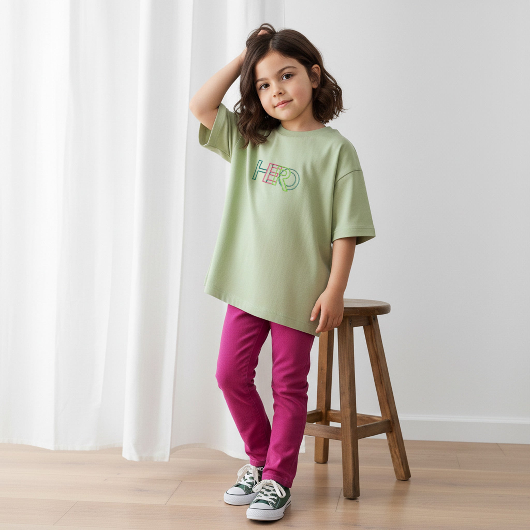 Fuşya rengi tayt ve haki yeşili, 'HERO' nakışlı oversize tişört giyen kız çocuk, elini saçına götürerek havalı bir poz veriyor.