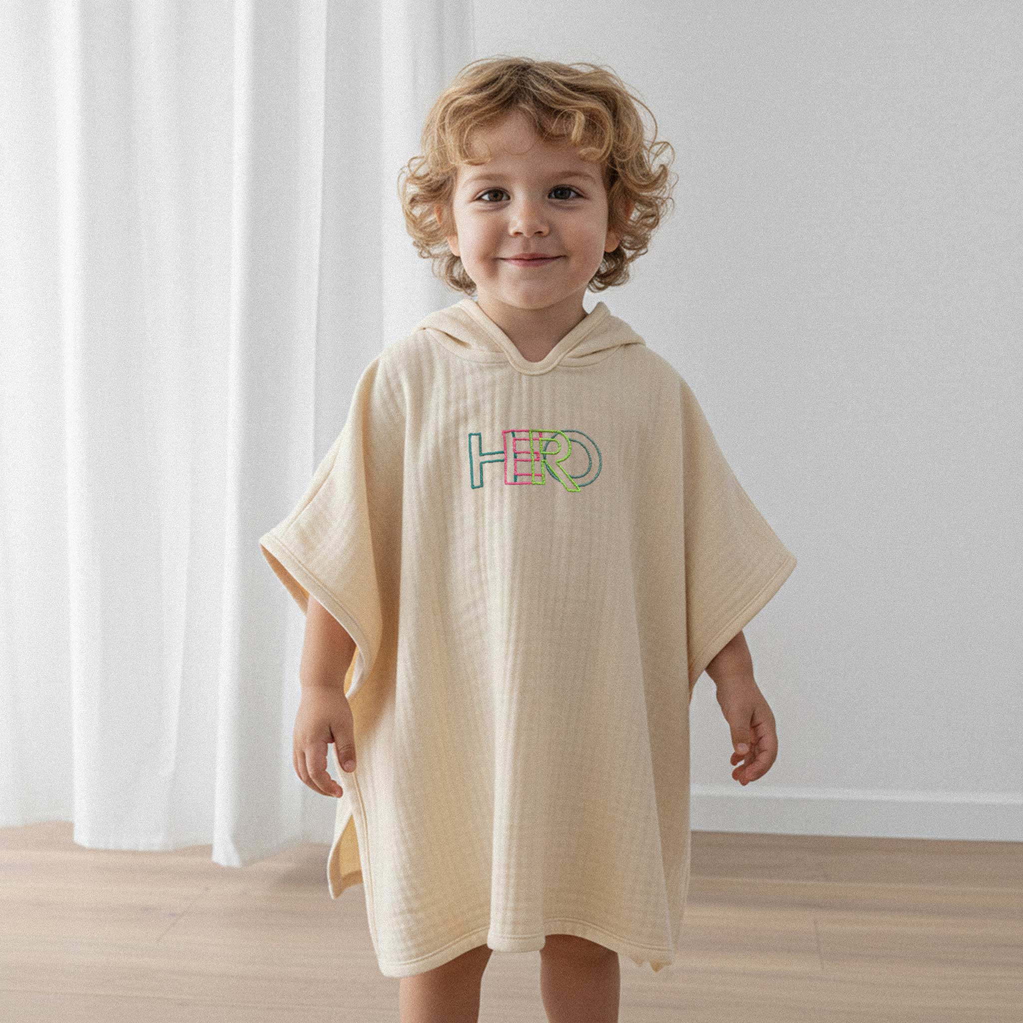 Kıvırcık saçlı küçük erkek çocuk, üzerinde ekru renkli, 'HERO' nakışlı kapüşonlu müslin panço ile sevimli bir şekilde gülümseyerek poz veriyor.