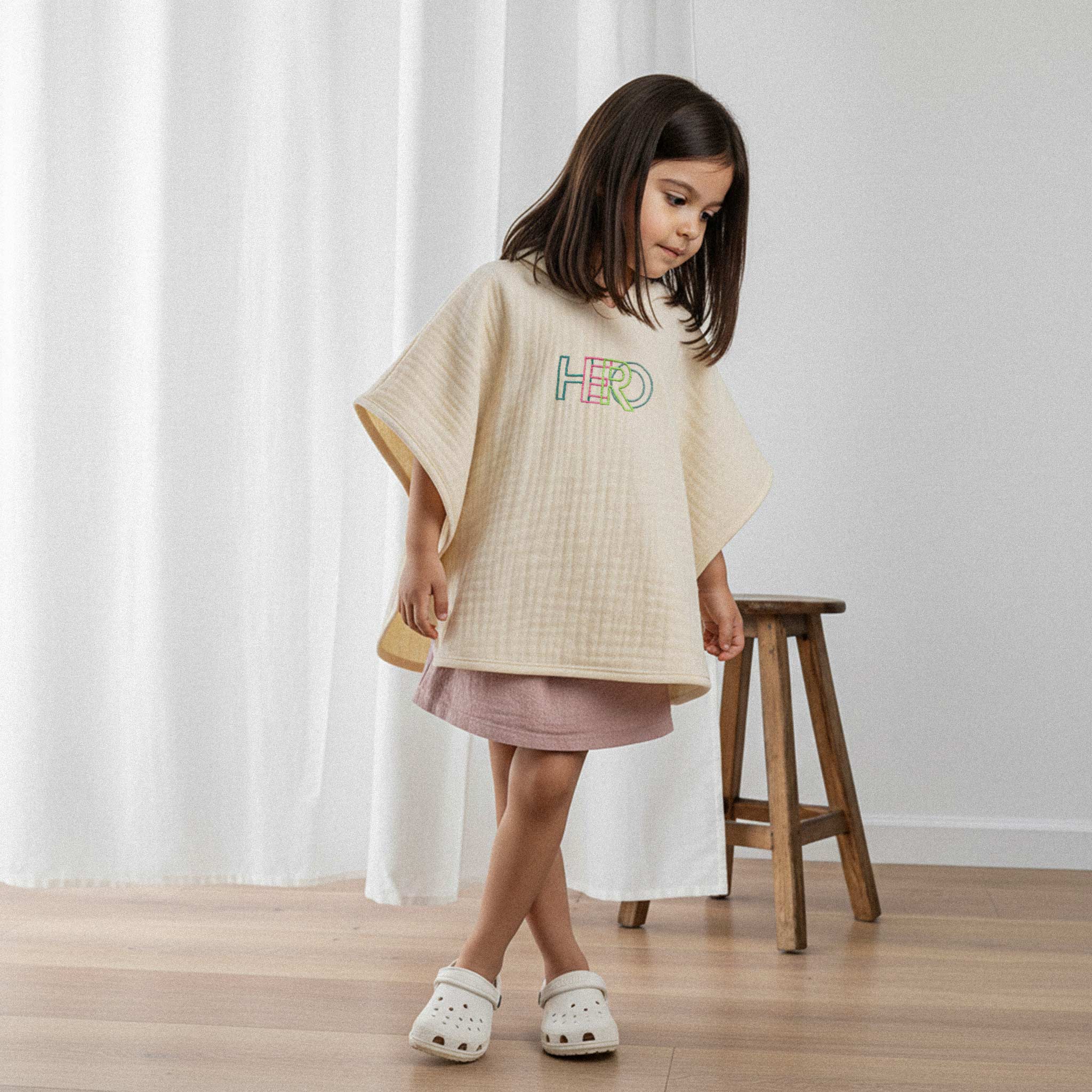 Pembe elbise üzerine ekru renkli, 'HERO' nakışlı kapüşonlu müslin panço giyen kız çocuk, şık bir şekilde poz vererek pançosuna bakıyor.