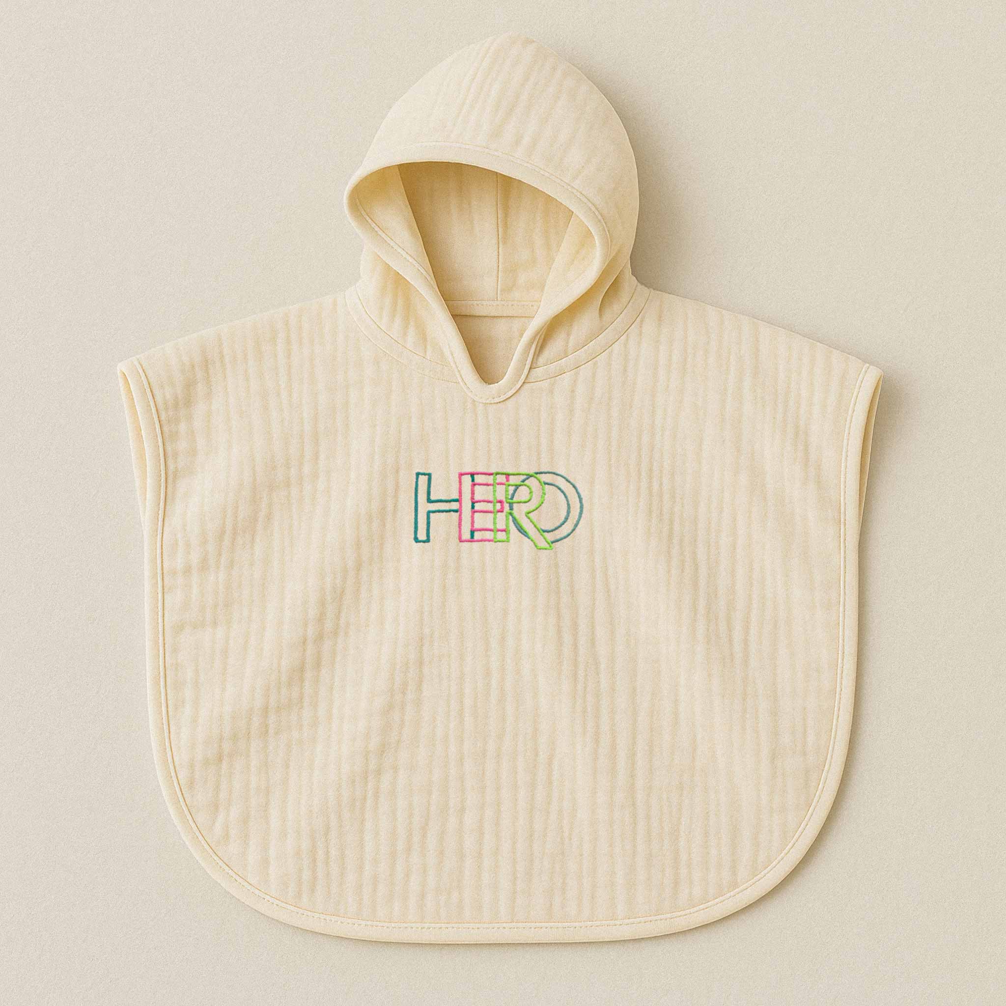 Ekru (doğal bej) renginde, kapüşonlu, müslin kumaştan bebek ve çocuk pançosu. Göğüs kısmında renkli ipliklerle "HERO" yazısı işlenmiştir.